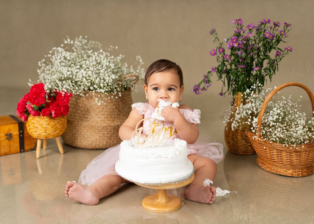 Baby in white dress explores first birthday cake surrounded by floral arrangements.