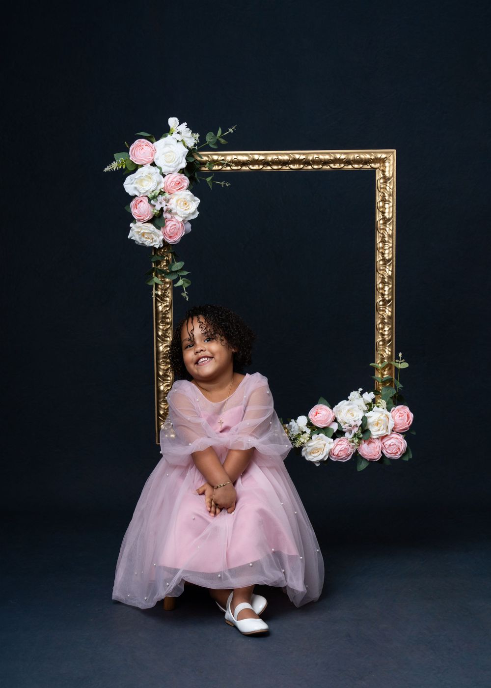 Child in pink tulle dress poses with ornate gold frame decorated with roses against dark backdrop.