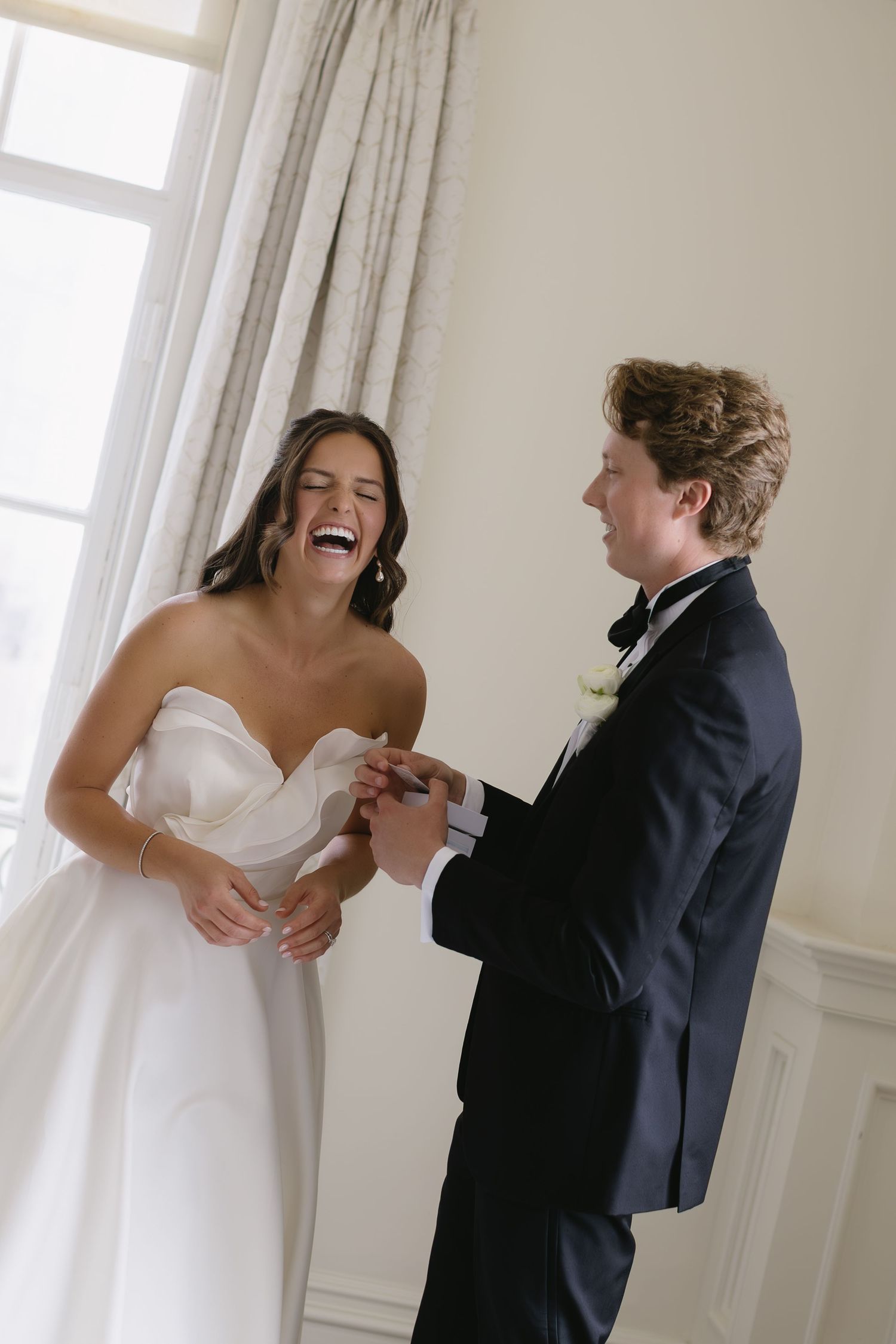 A joyful wedding moment as a bride in white strapless gown laughs while exchanging rings with groom in classic tuxedo.