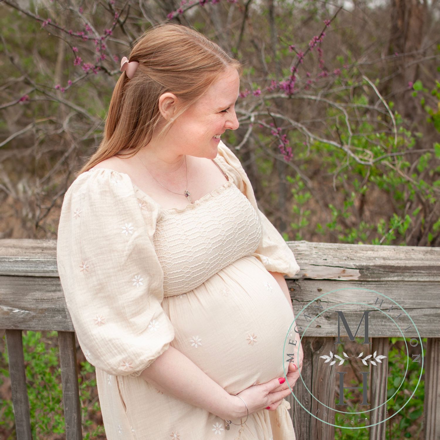 A pregnant mama in a white flowing dress stands at a rustic wooden fence surrounded by blooming spring foliage.