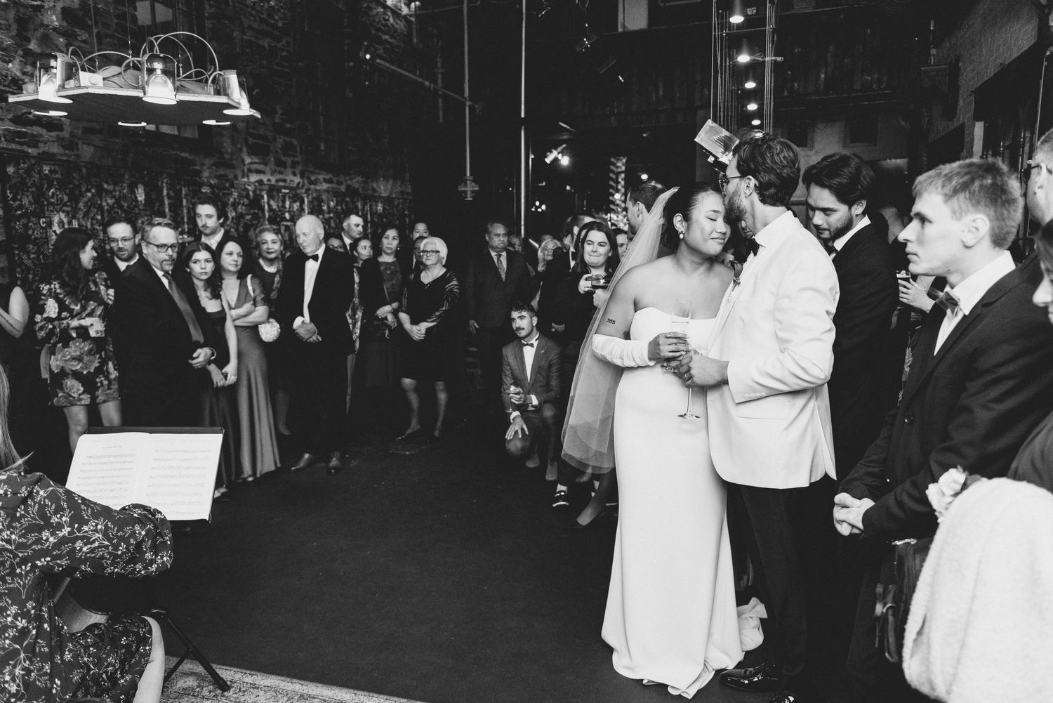 A black and white wedding photograph showing a couple sharing their first dance surrounded by guests at their reception.
