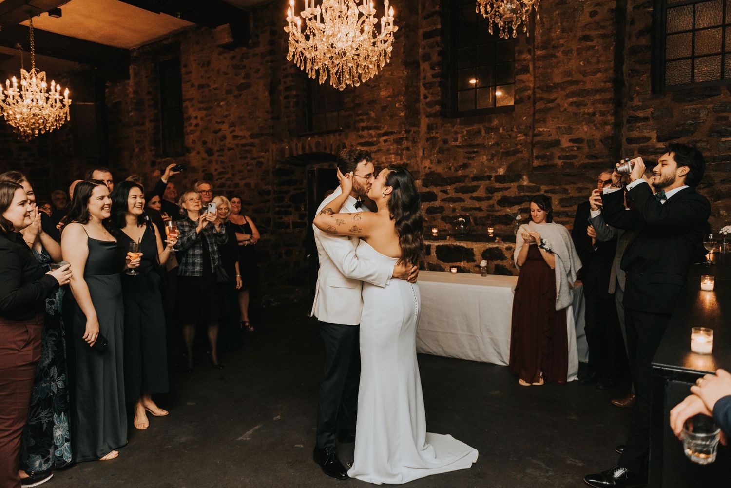 Two people share an intimate dance while guests in dark formal attire look on in a rustic brick venue with crystal chandeliers.