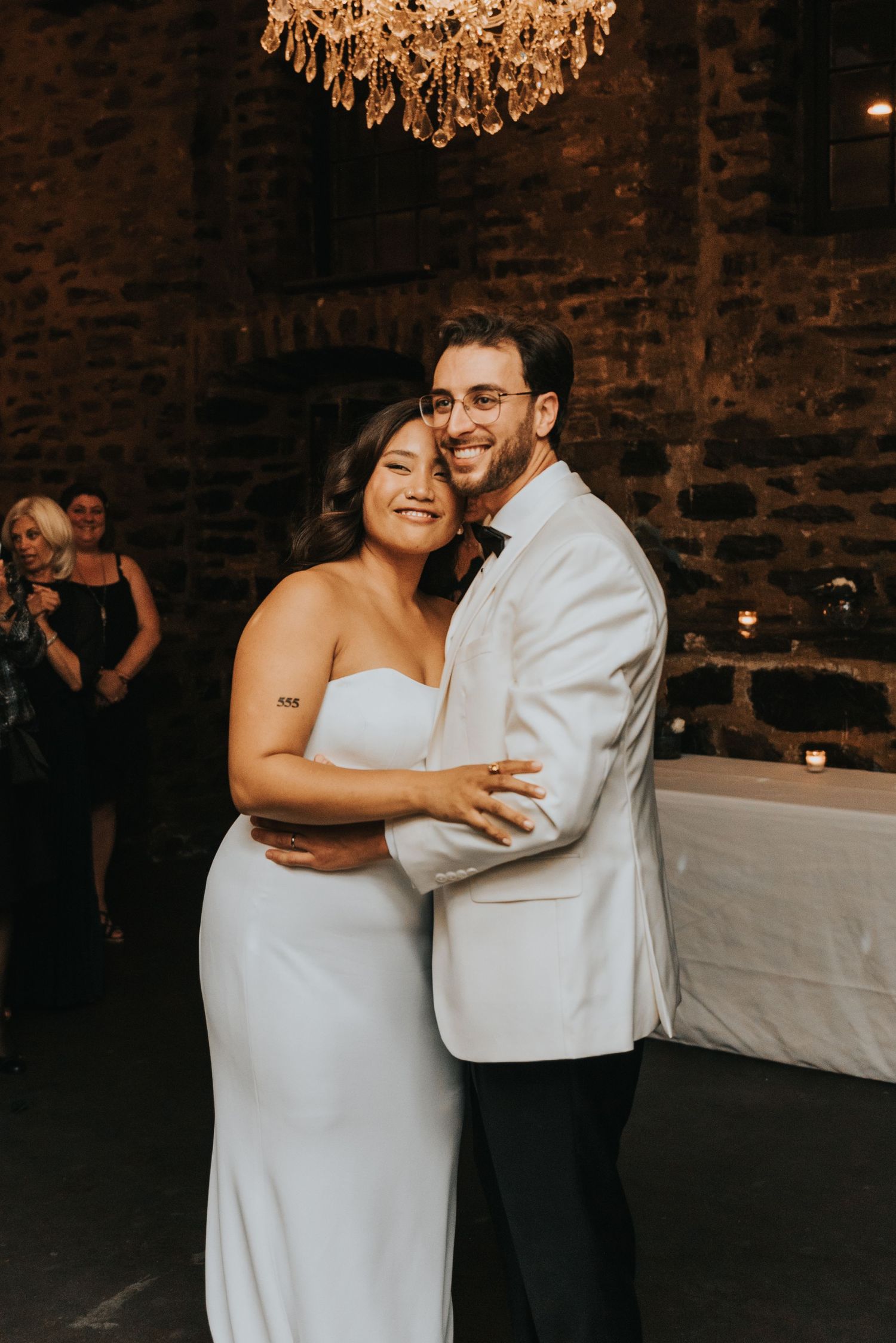 Two people share a joyful embrace on their wedding day in a rustic brick venue under an elegant crystal chandelier.