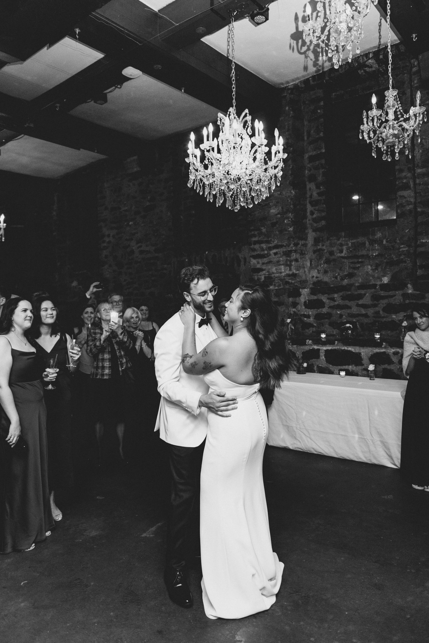 A black and white photo captures a romantic first dance moment at a wedding reception under crystal chandeliers.