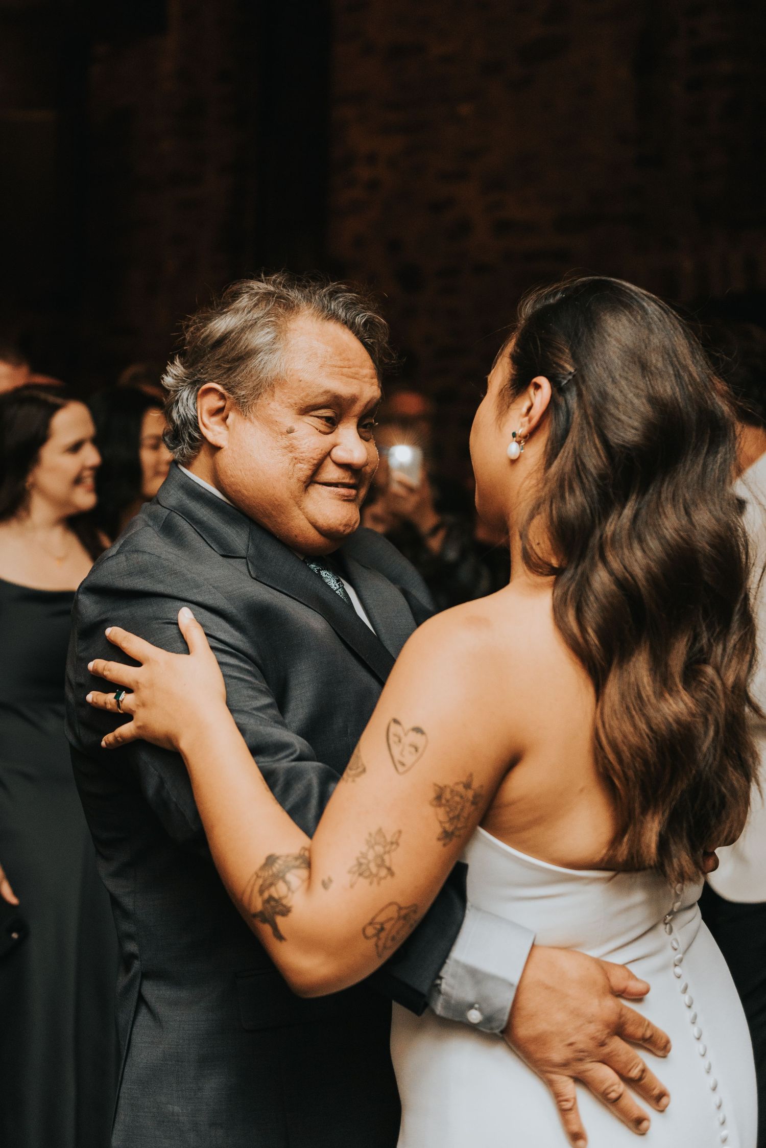 Two people share an emotional moment dancing together at what appears to be a wedding reception in a dark venue.