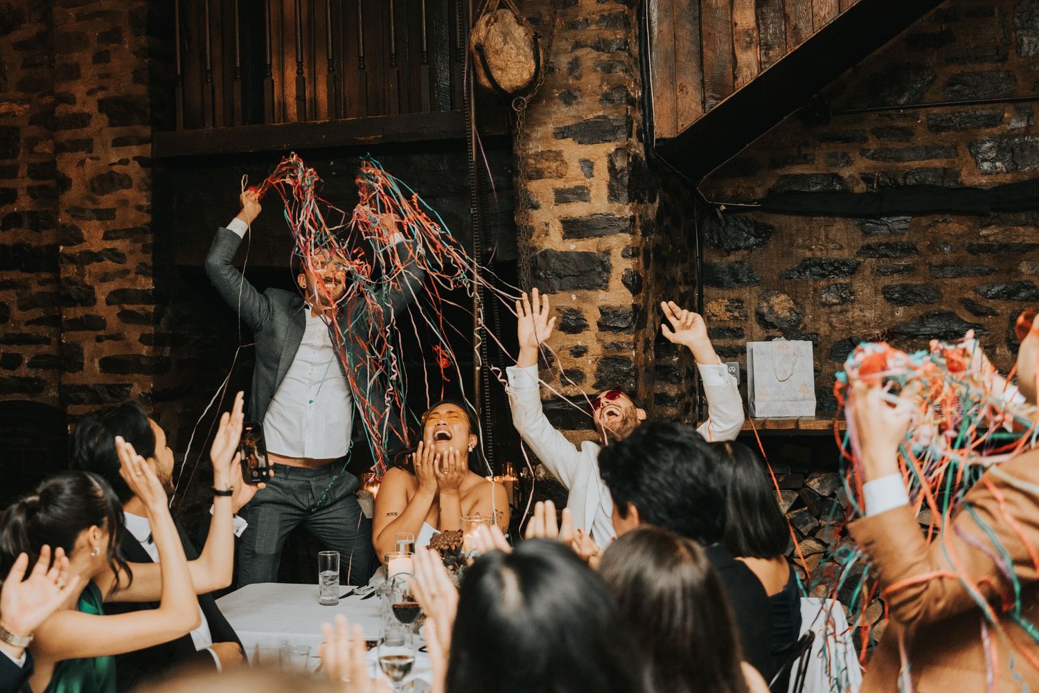 Celebrating crowd throws confetti in the air during a festive party moment at rustic brick venue.