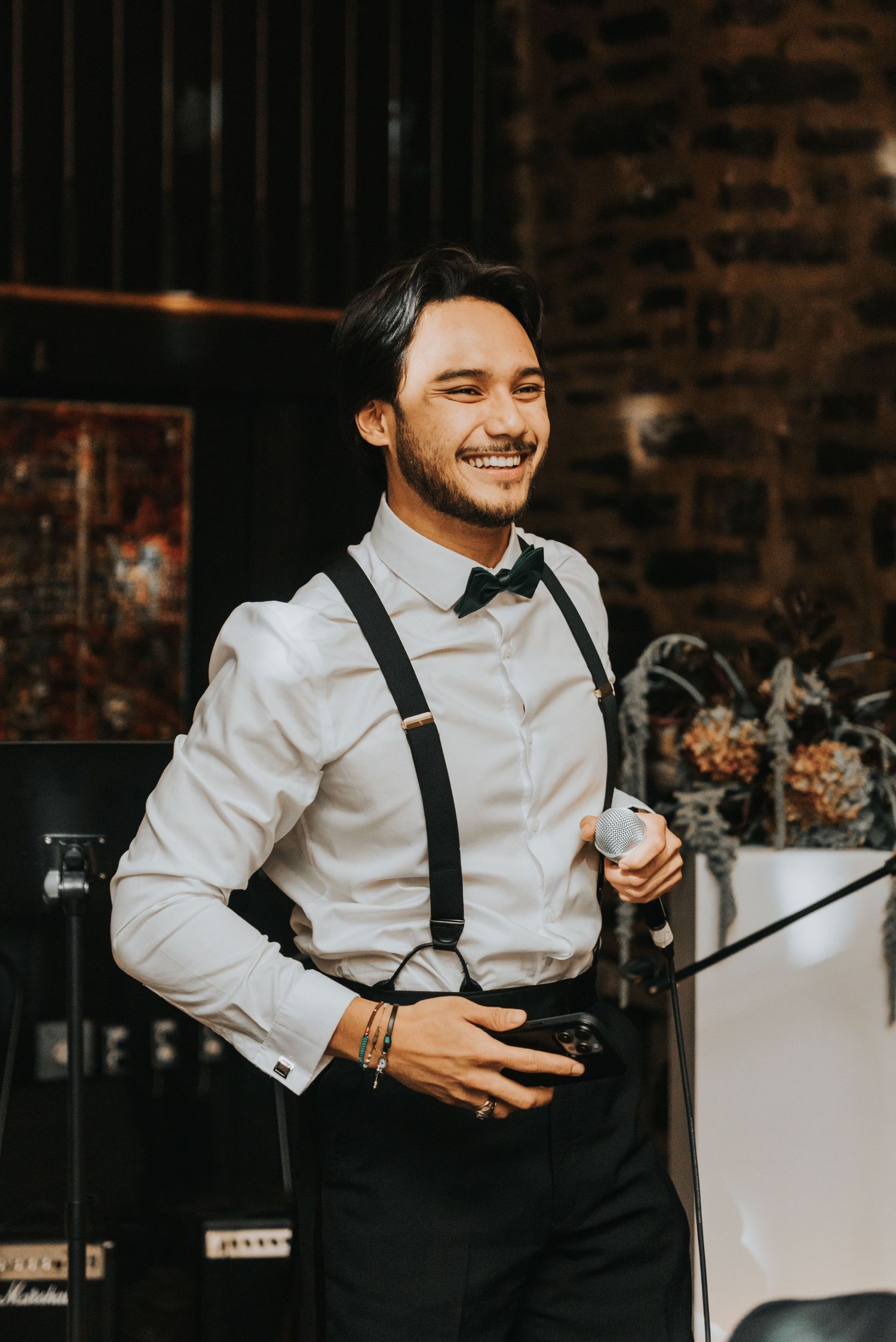 Professional photographer in classic white shirt and suspenders smiles warmly while holding camera at an event.