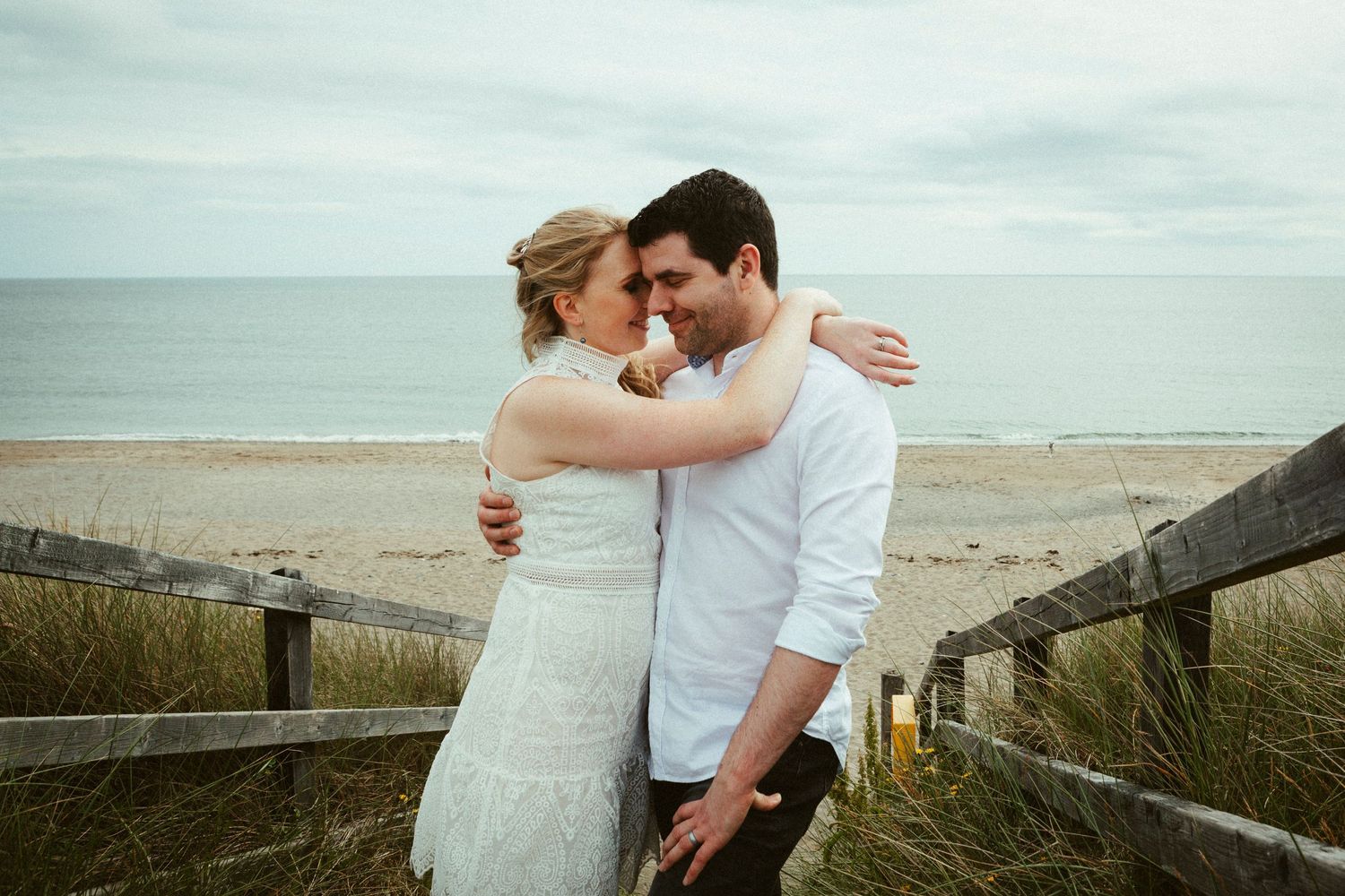 A couple embraces on a wooden boardwalk overlooking a scenic beach under an overcast sky.