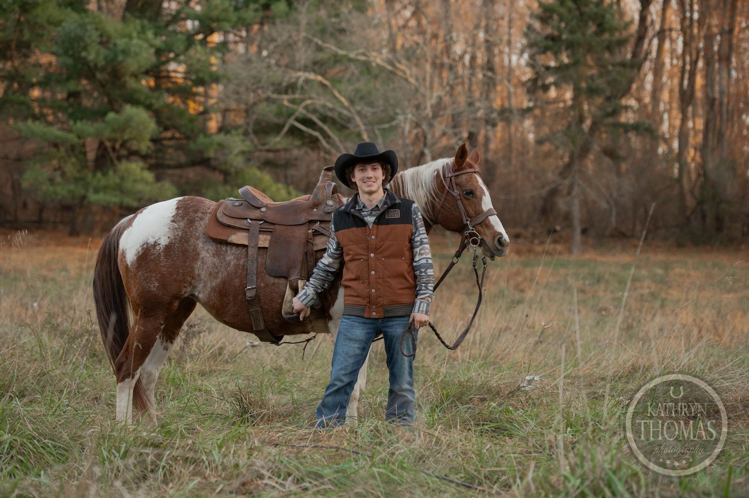A paint horse with western tack stands with a rider in a brown vest and black hat in a fall field.