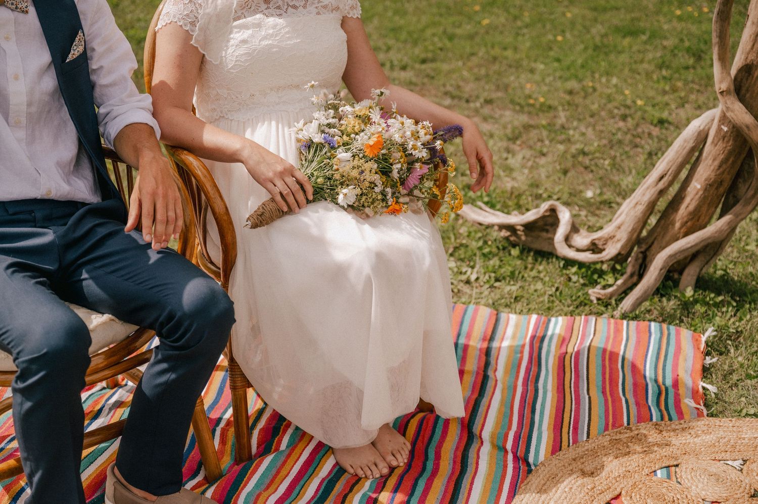 Wedding couple sits on striped blanket holding rustic bouquet during outdoor celebration.