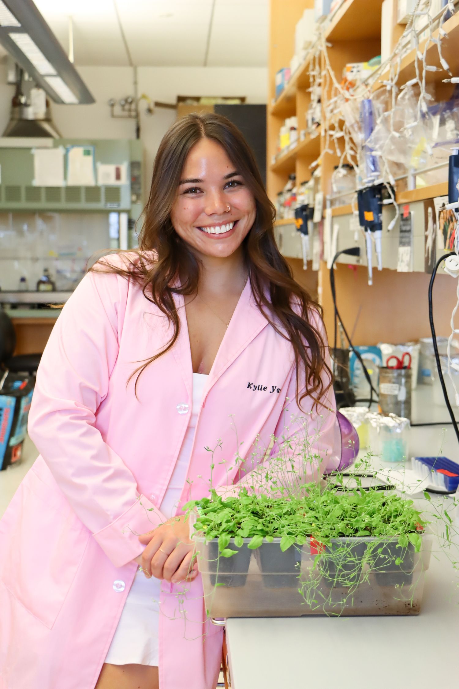 Student in pink lab coat works with plant specimens in research laboratory.
