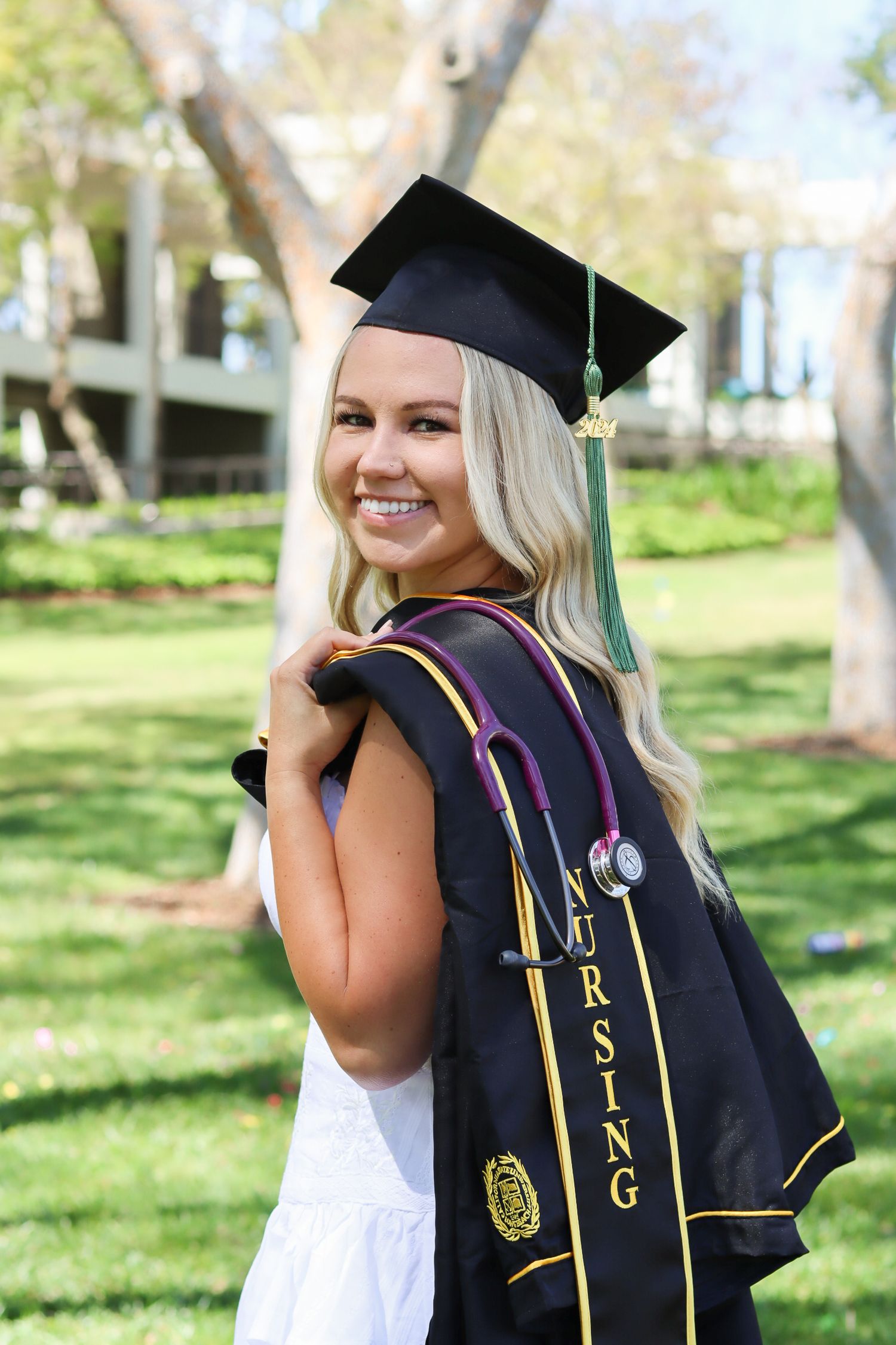 Graduate in white dress and black stoles poses for graduation photos in park setting.