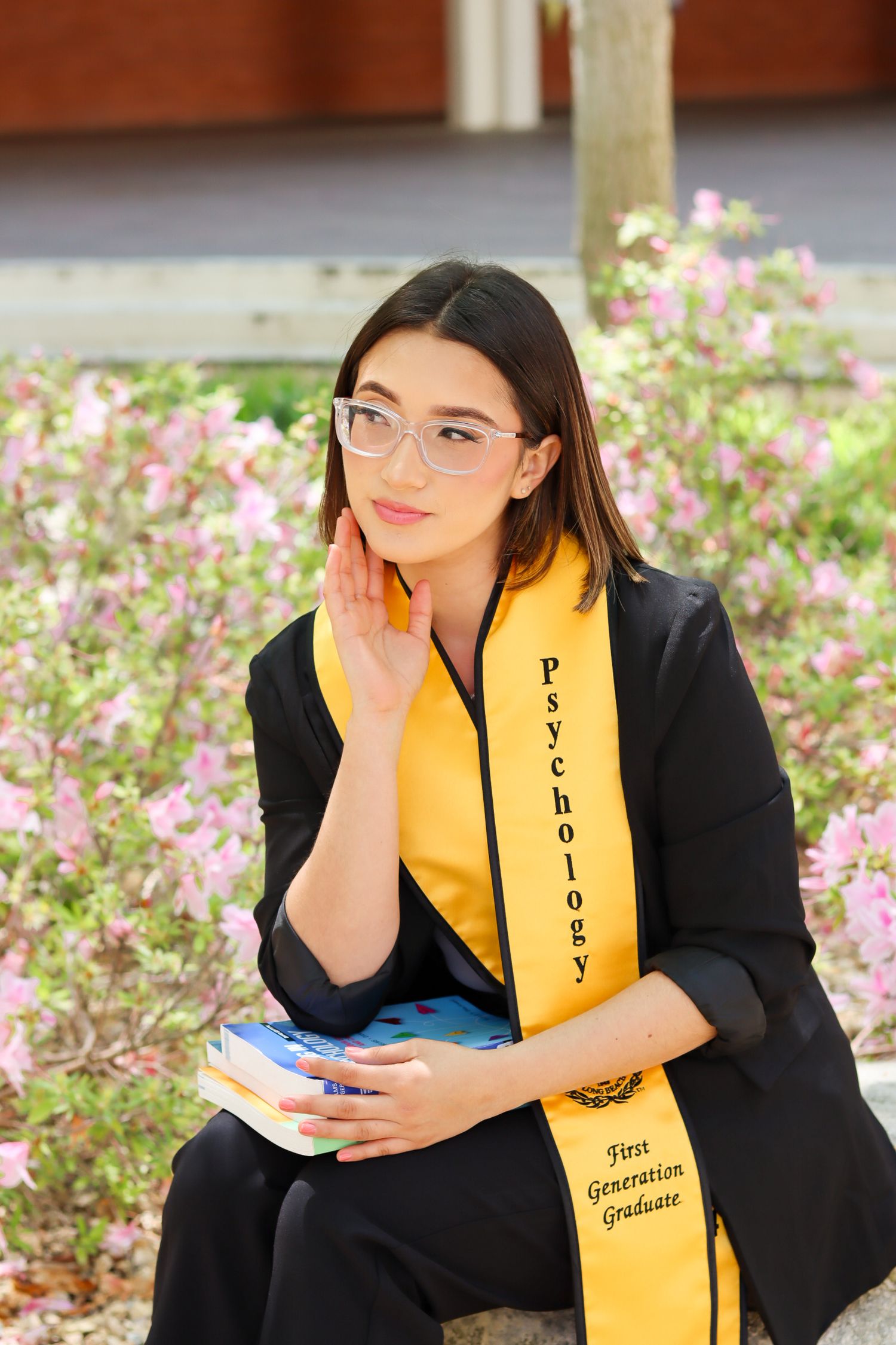 Graduate in black gown and yellow stole poses near pink flowers.