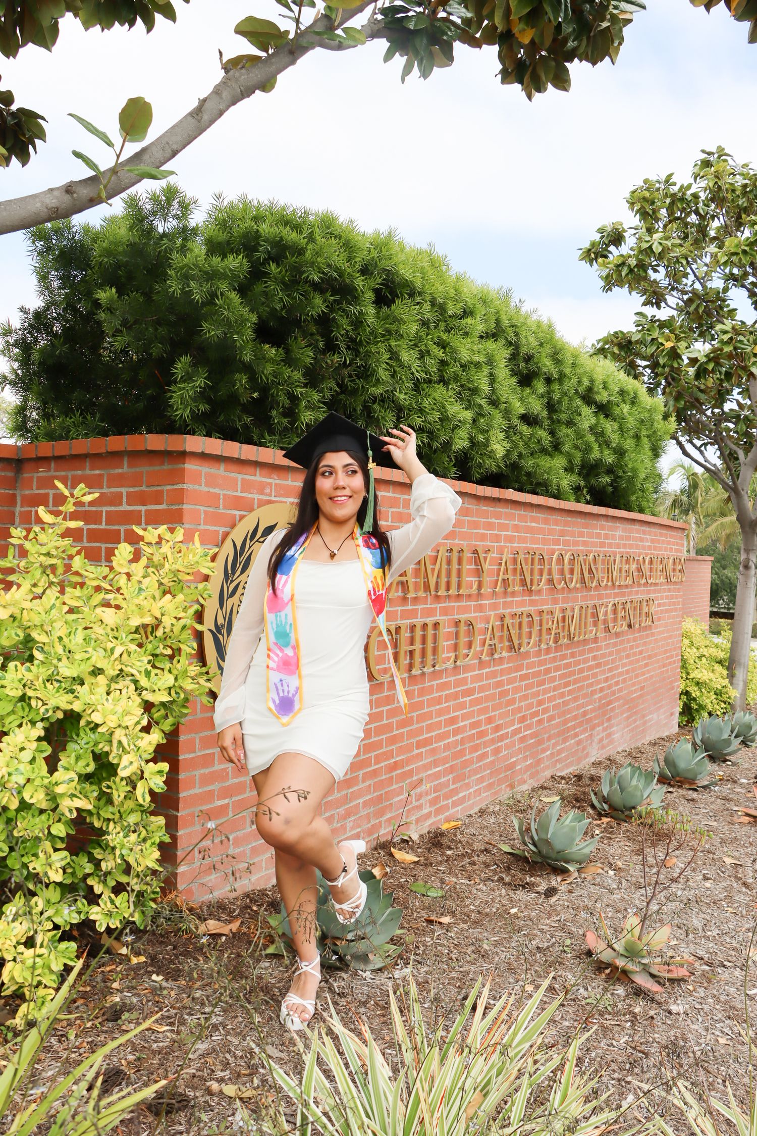 Graduate takes photos in front of brick campus sign surrounded by landscaping.