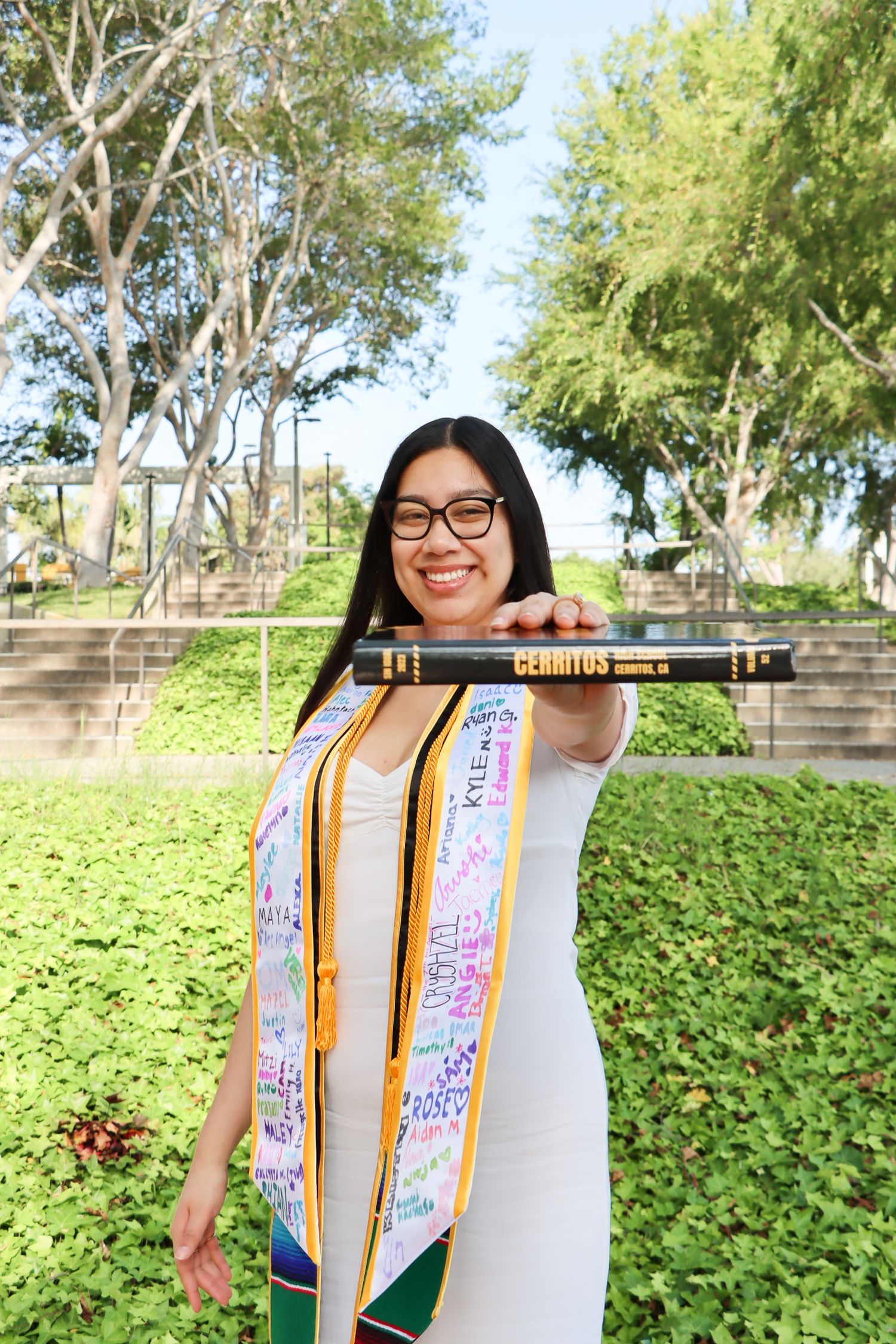 Graduate wearing decorated sash poses in front of green campus landscaping.