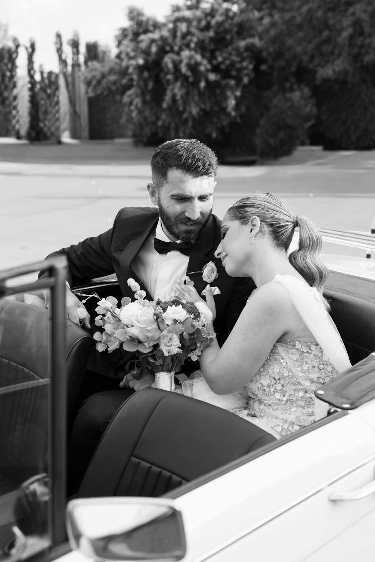 A groom in a light grey suit with a white bow tie reads a document while smiling outdoors.