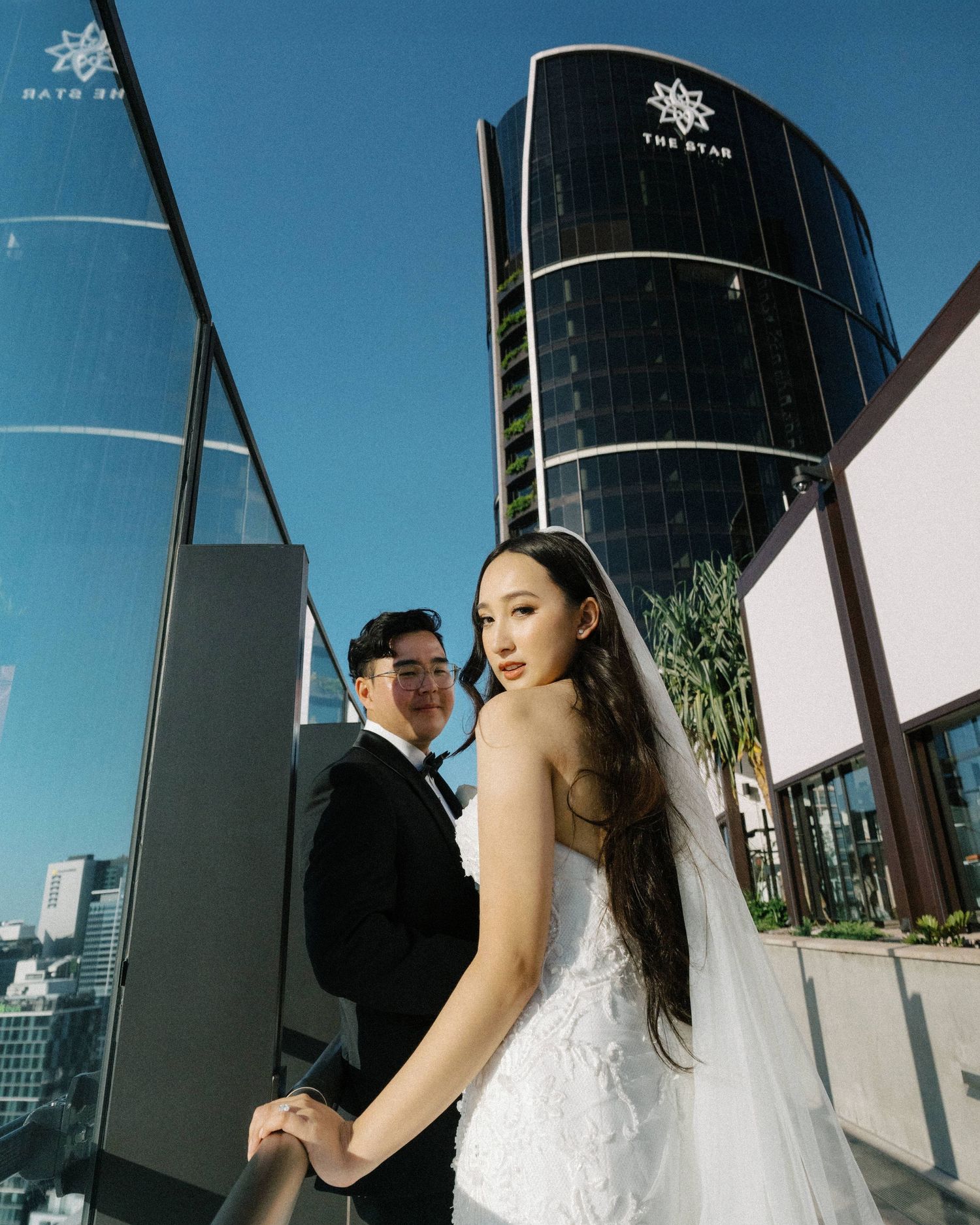 A couple in wedding attire poses on a rooftop terrace with modern glass buildings and blue sky in the background.