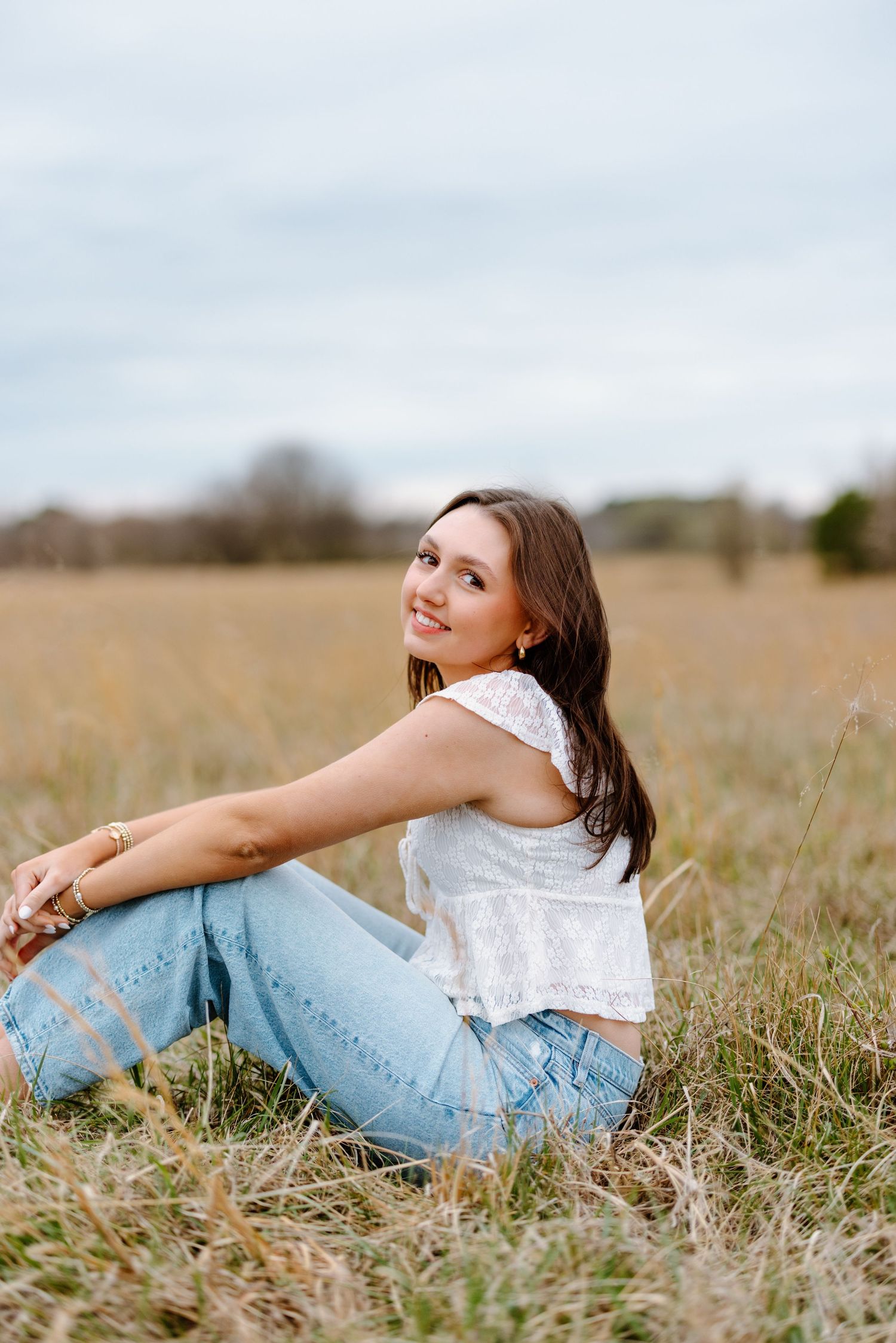 Young woman in white top and jeans sits relaxed in tall grass field on a peaceful autumn day.