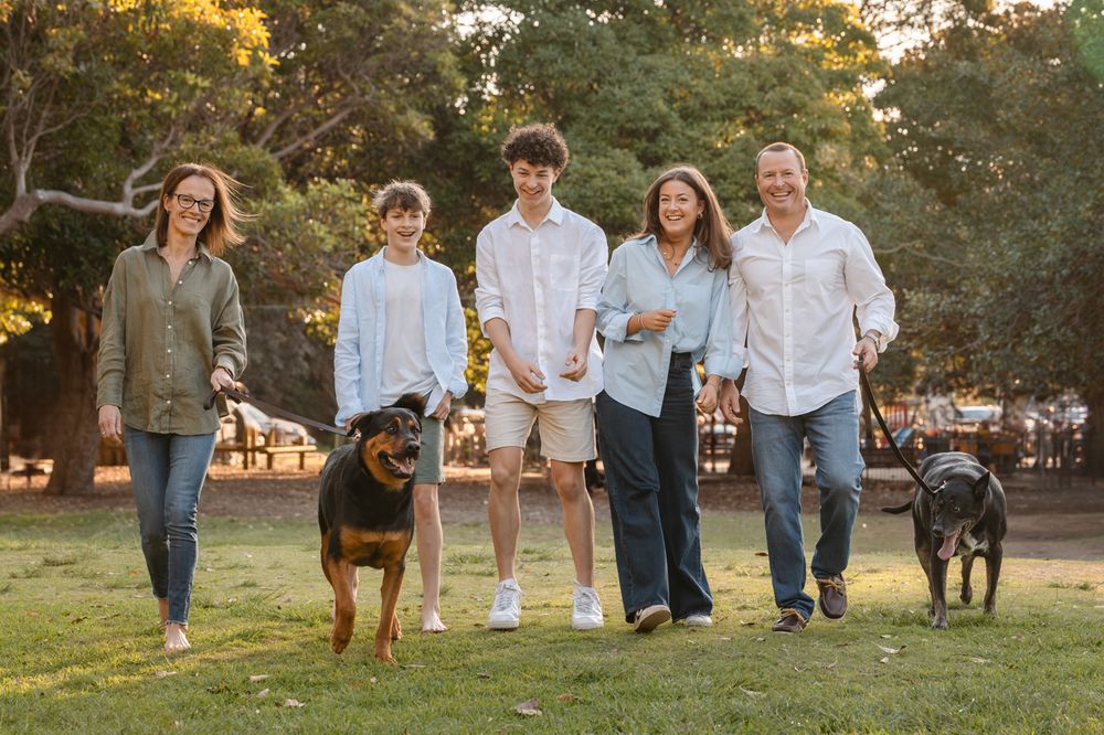 Family of four walks with two dogs in a sunny park, smiling and enjoying quality time together outdoors in mosman family photoshoot