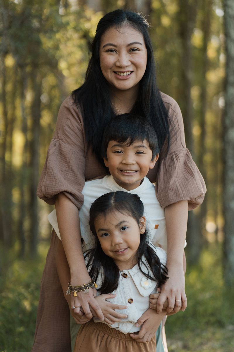 A smiling Asian mother hugs her two young children outdoors in a forest setting in rouse hill regional park family photoshoot
