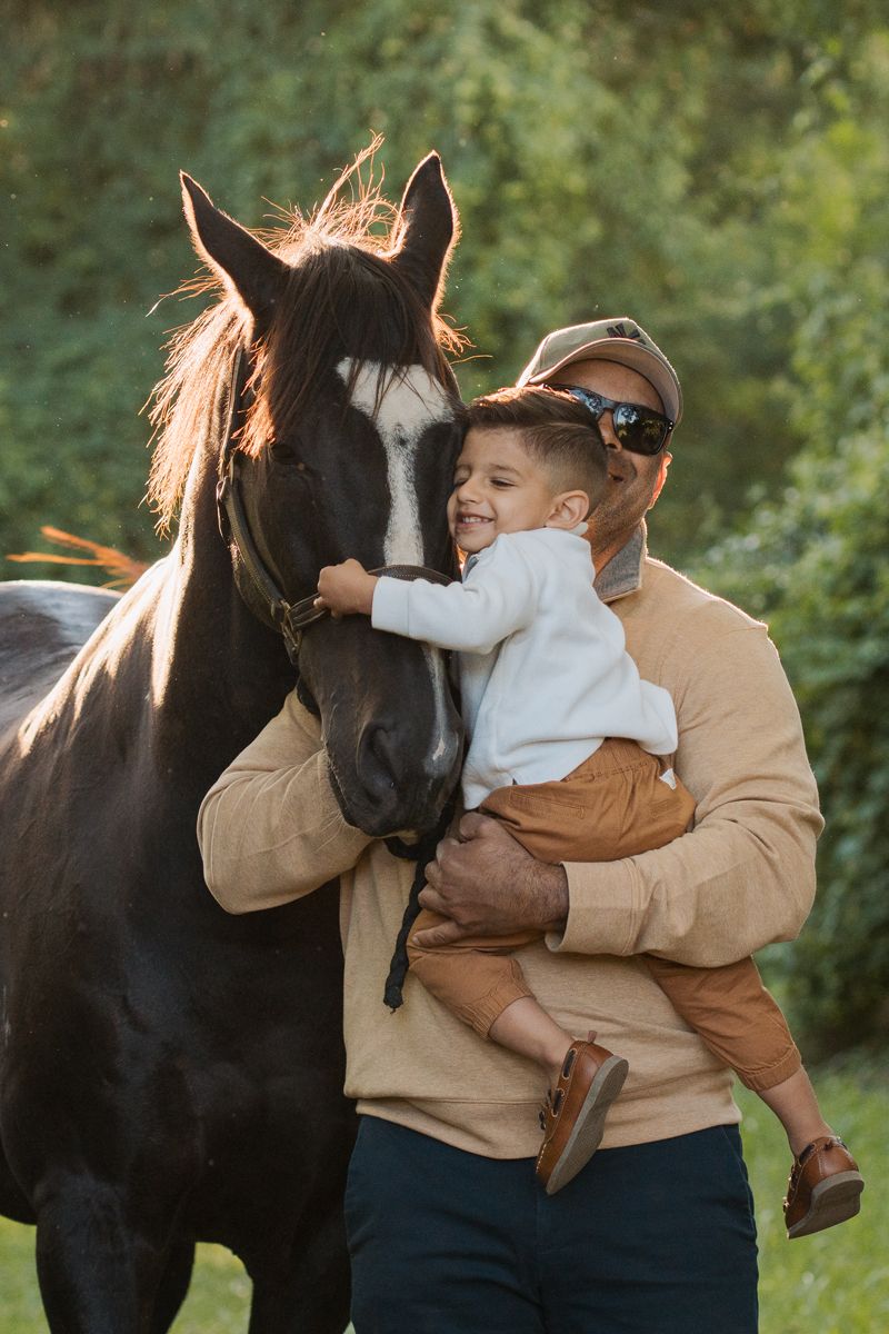 Man holding young child who hugs a dark horse outdoors in golden sunlight near green trees in Pitt town horse photoshoot with a family