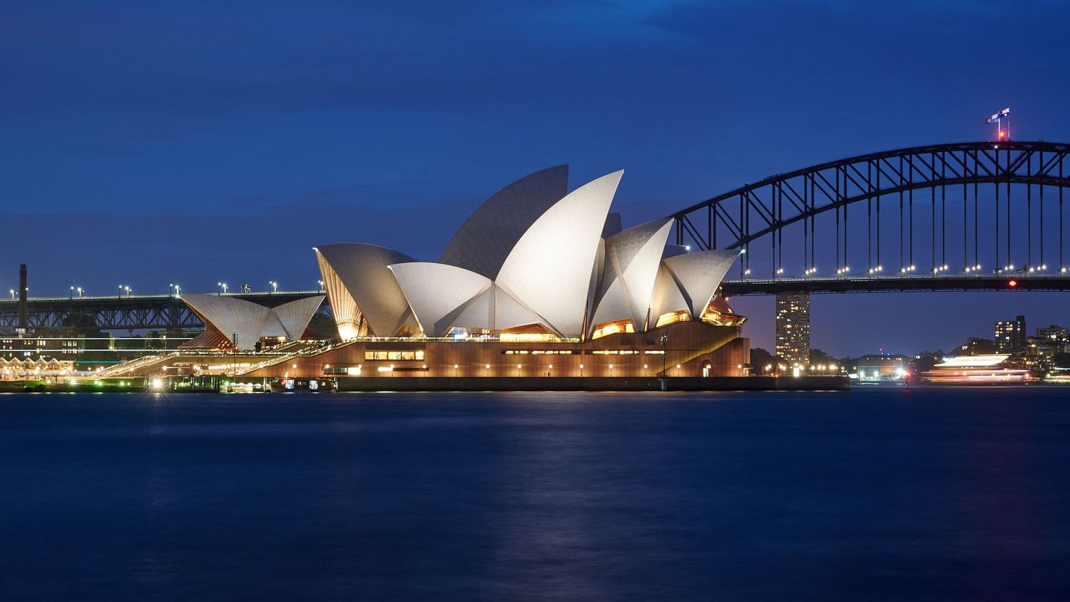 The Sydney Opera House and Harbour Bridge illuminate the night skyline with stunning reflections on the water.