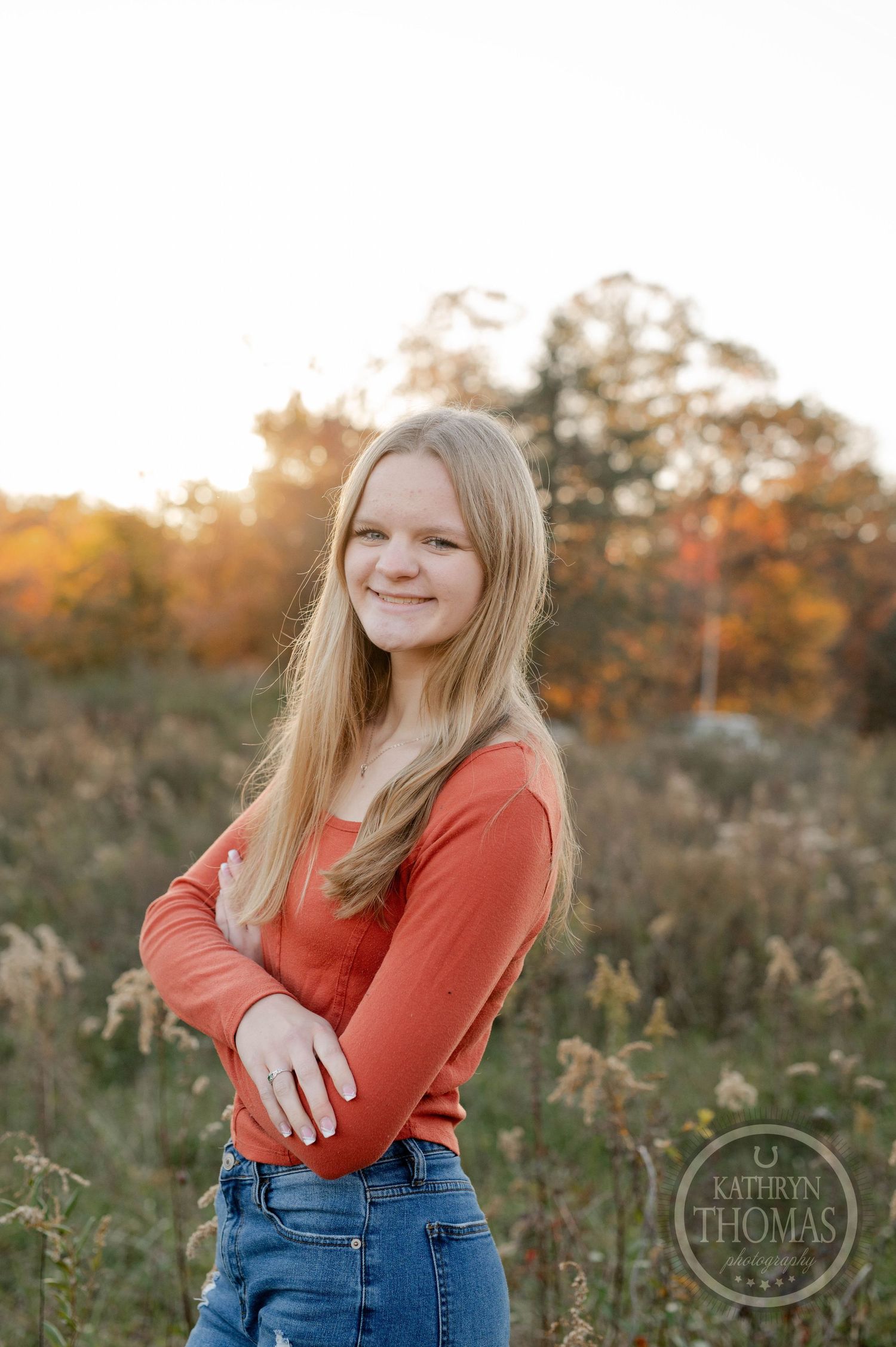 Person in coral sweater and jeans poses outdoors with autumn trees in soft lighting.