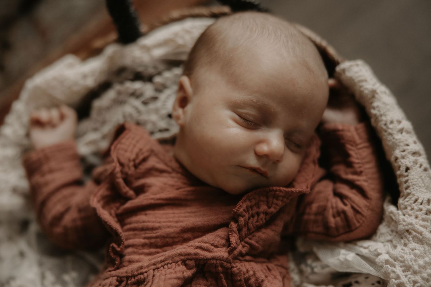 A newborn baby swaddled in a rustic brown wrap lies peacefully against a cream-colored textured blanket.