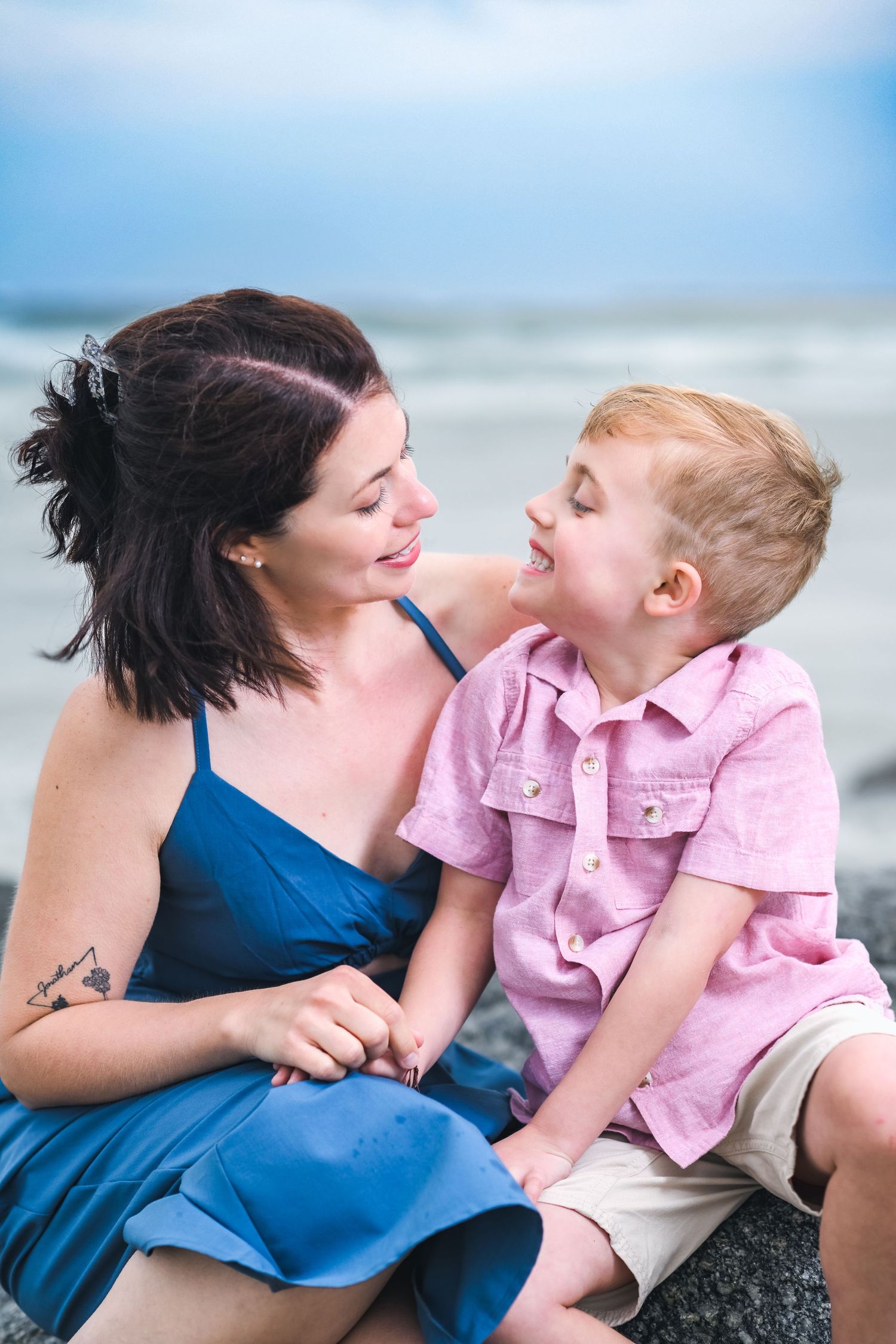 A tender moment shared between parent and child sitting on a rocky beach as they laugh together in blue and pink outfits.