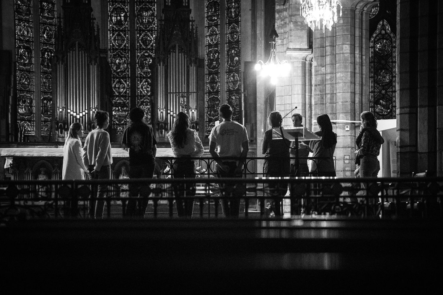 Photo en noir et blanc d'un chœur chantant dans le sein d'une église éclairée par des lustres. On aperçoit un orgue et des murs ornementés.