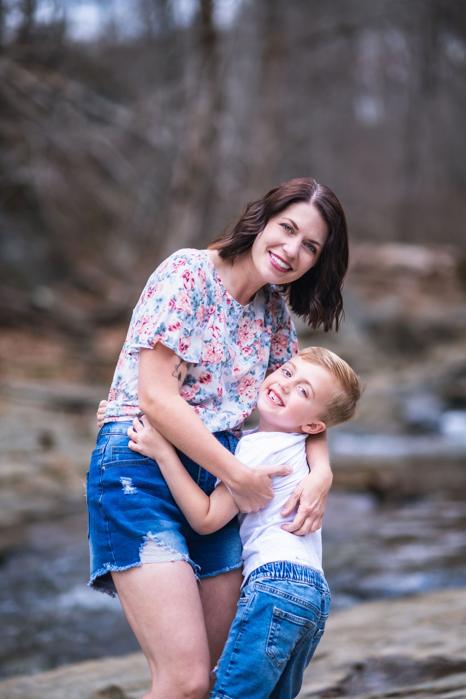 A tender moment between parent and child embracing outdoors by a creek in casual summer attire.