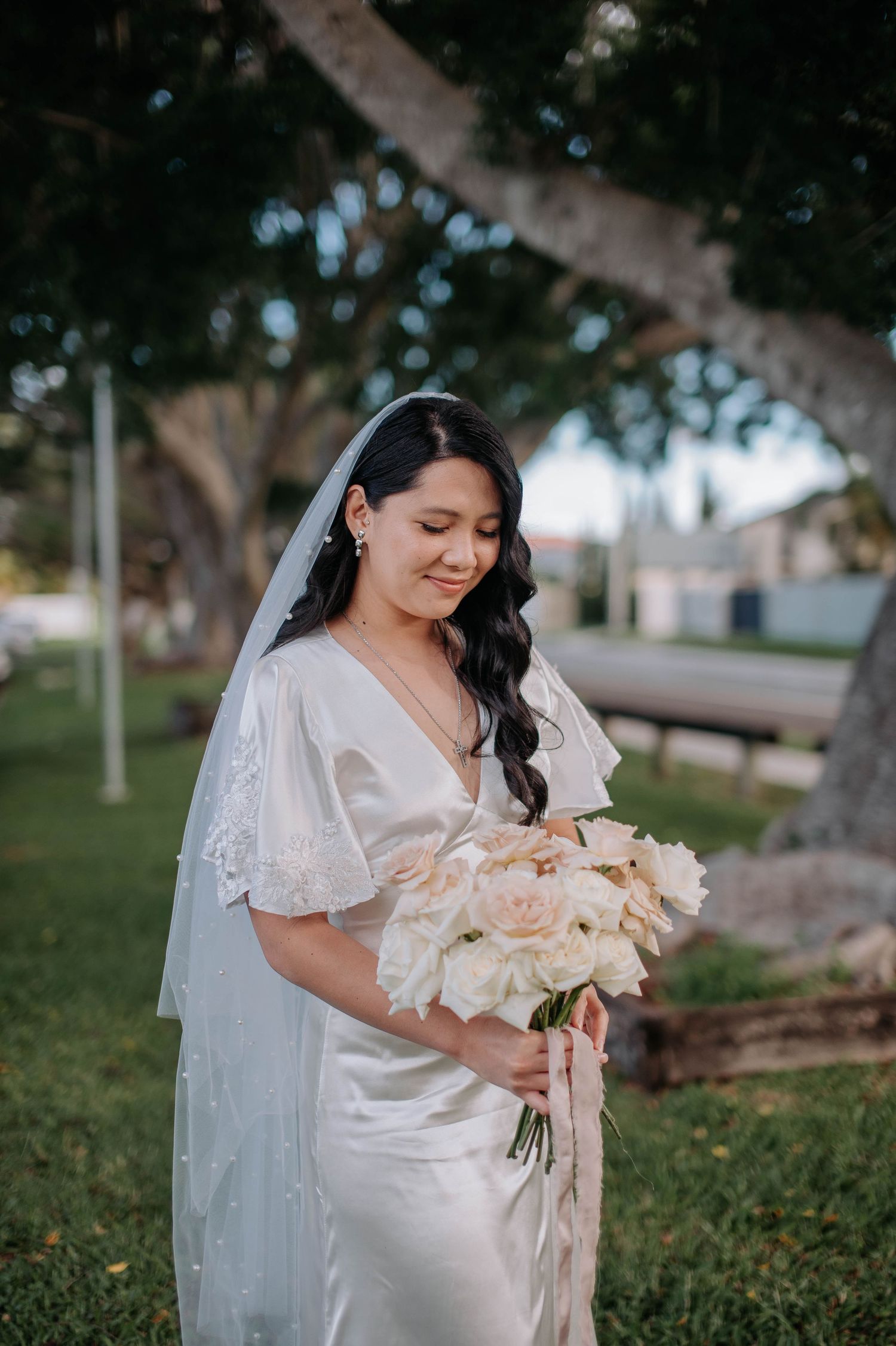 A bride in a flowing white dress holds a soft peach rose bouquet while standing in a garden setting.