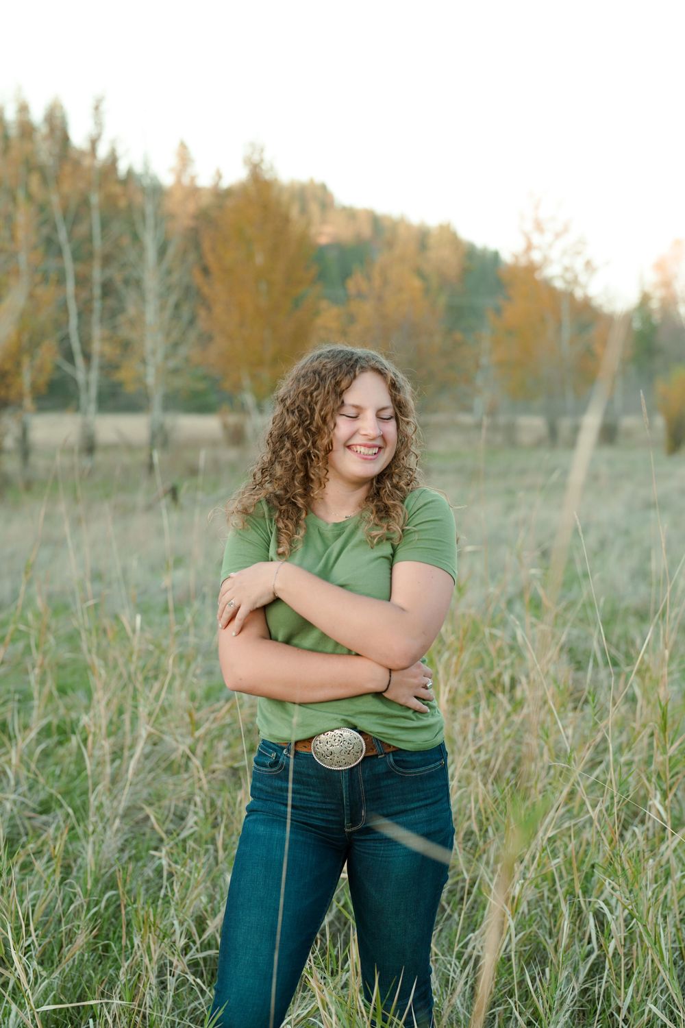 A person in a green shirt and jeans stands smiling in a field of tall grass during golden hour.