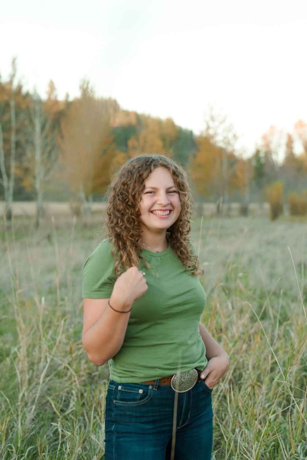 A person in a green t-shirt and jeans stands smiling in a field during golden hour with autumn trees in background.