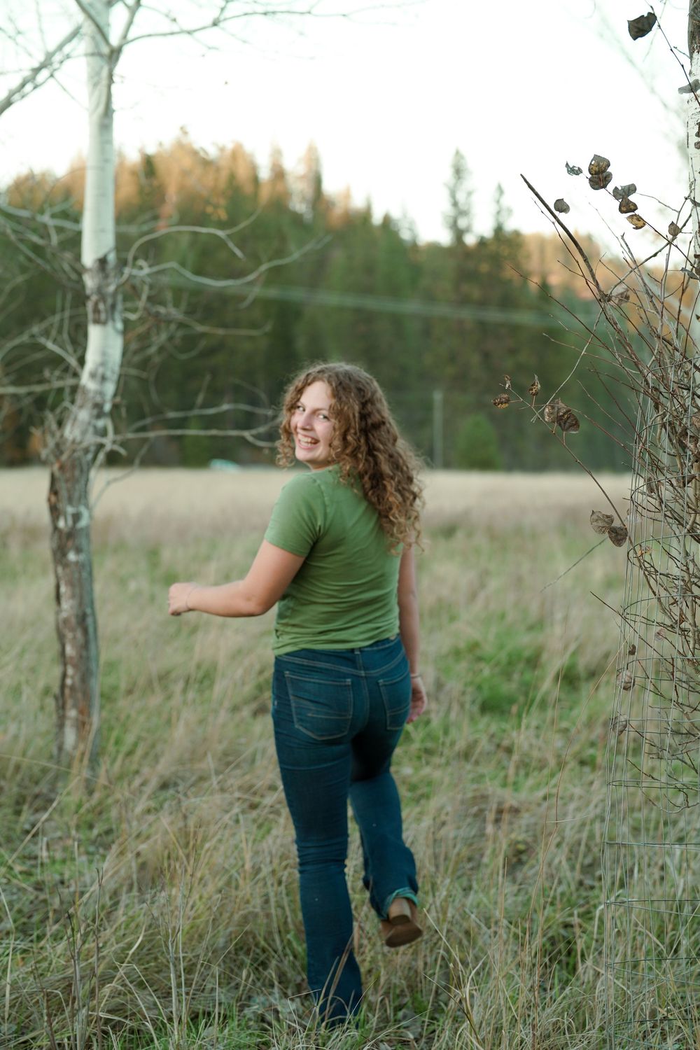 Person in a green shirt and jeans stands in a grassy field at sunset with autumn trees in the distance.