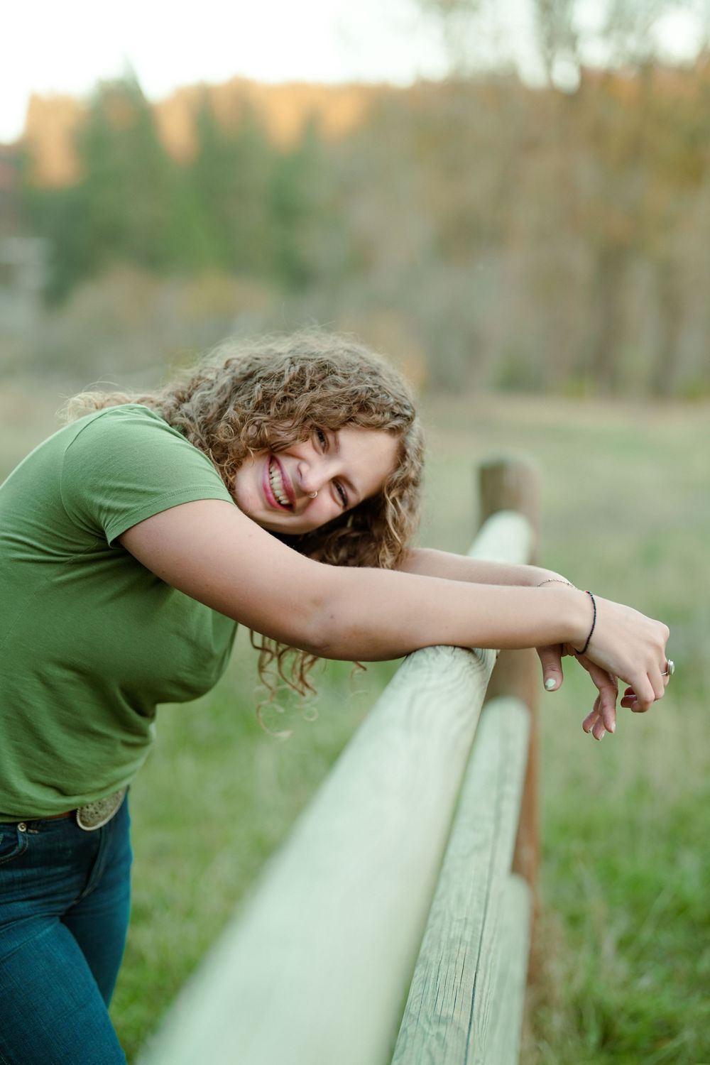 Person in green shirt leaning on wooden fence rail outdoors with natural blurred background in sunlight.