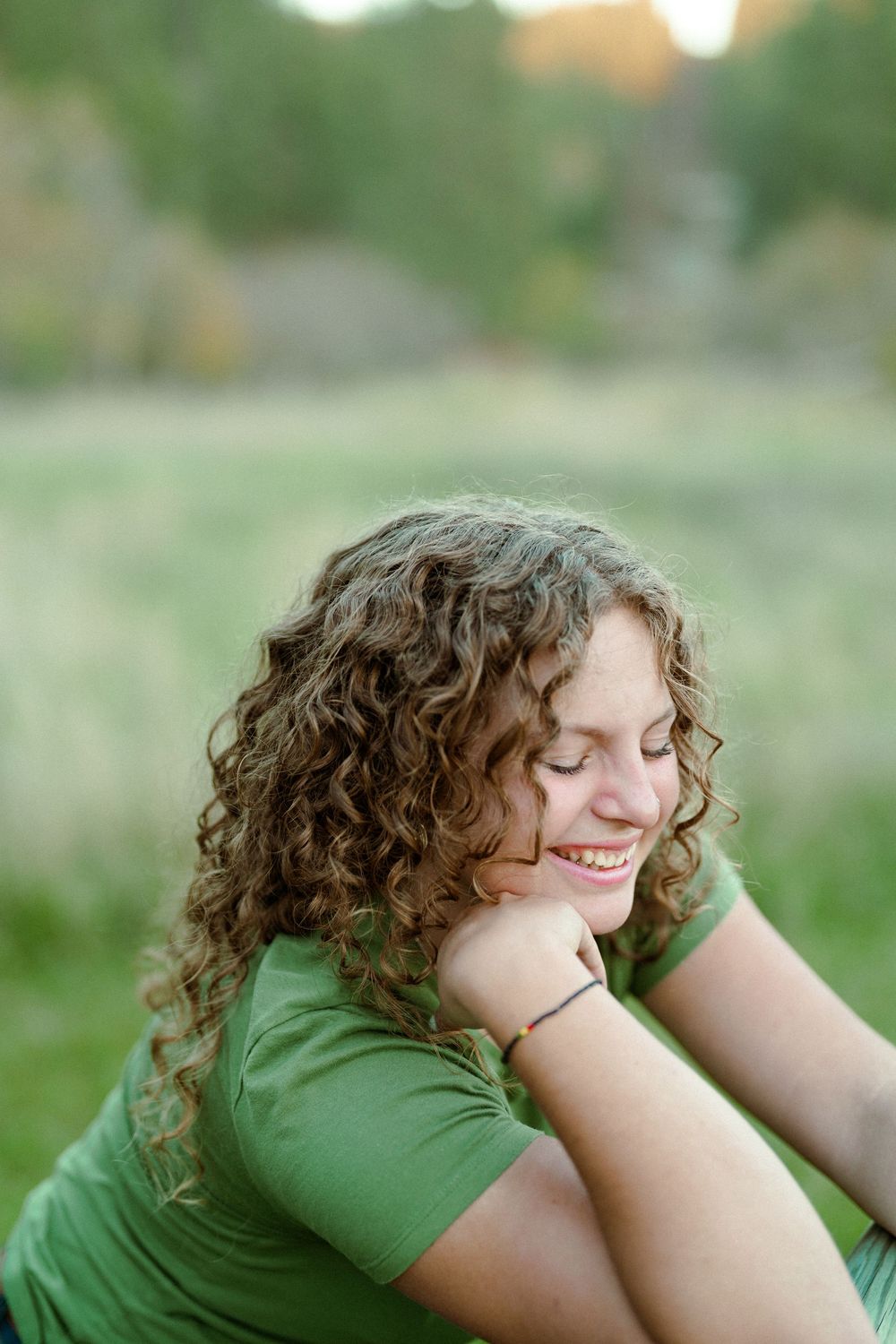 A person with curly hair and a green shirt sits outdoors with a joyful expression on a sunny day.