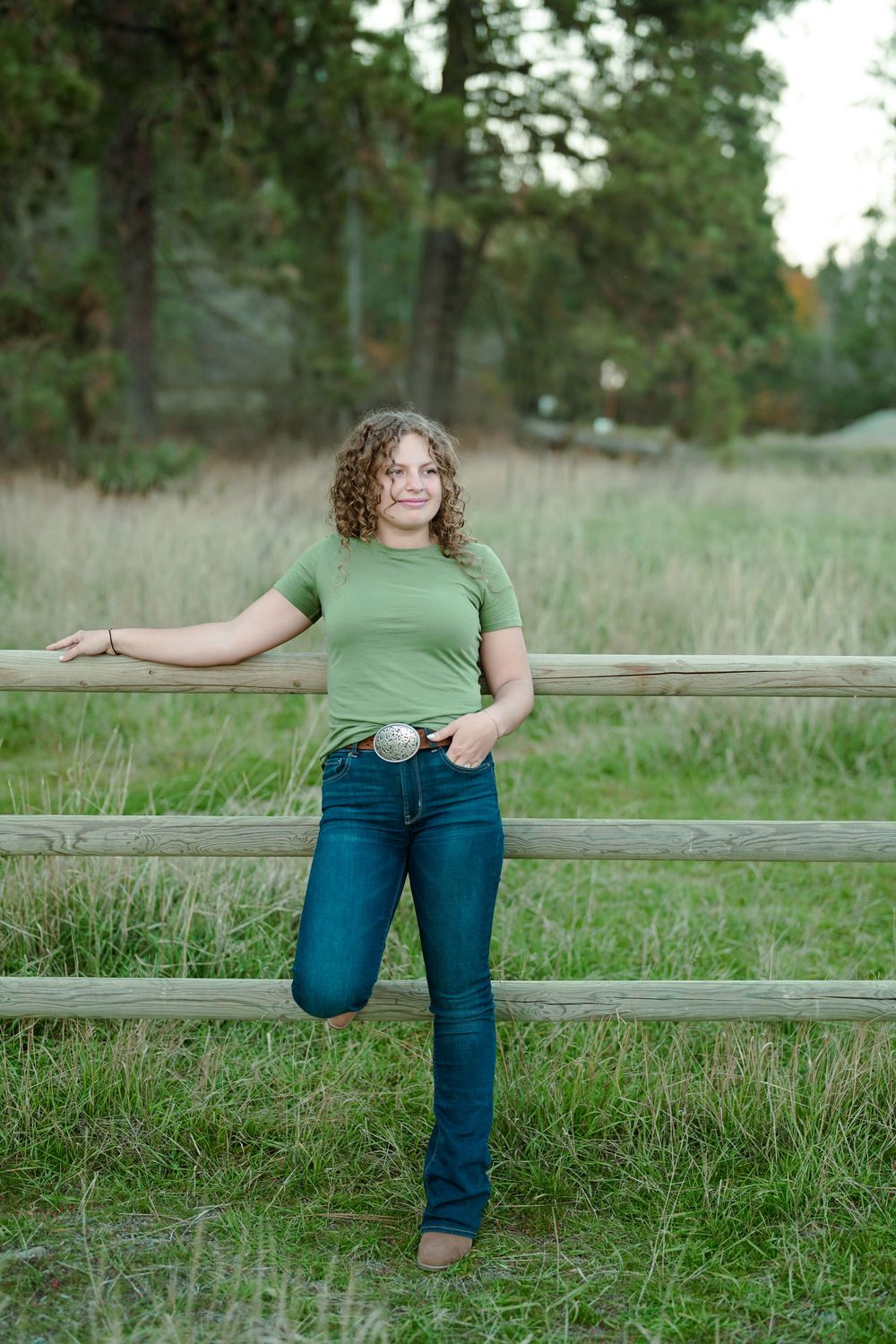 A casual portrait of someone leaning against a rustic wooden fence in a grassy field wearing jeans and green t-shirt.