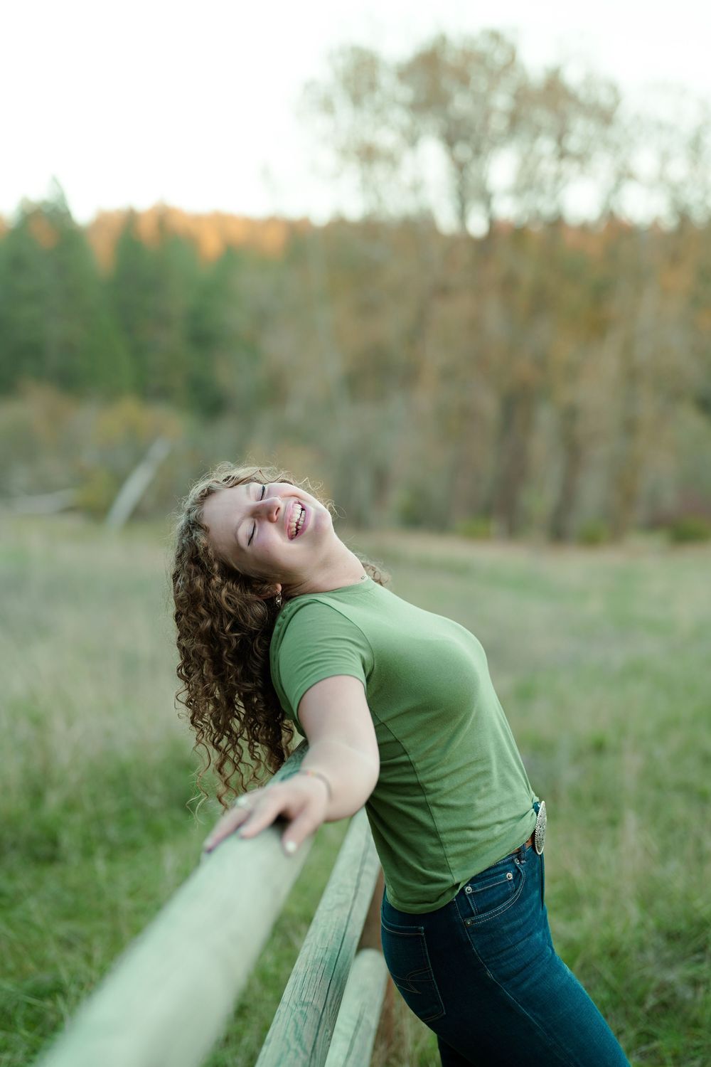 Person in a green t-shirt and jeans leans back playfully against a wooden fence in an autumn field.