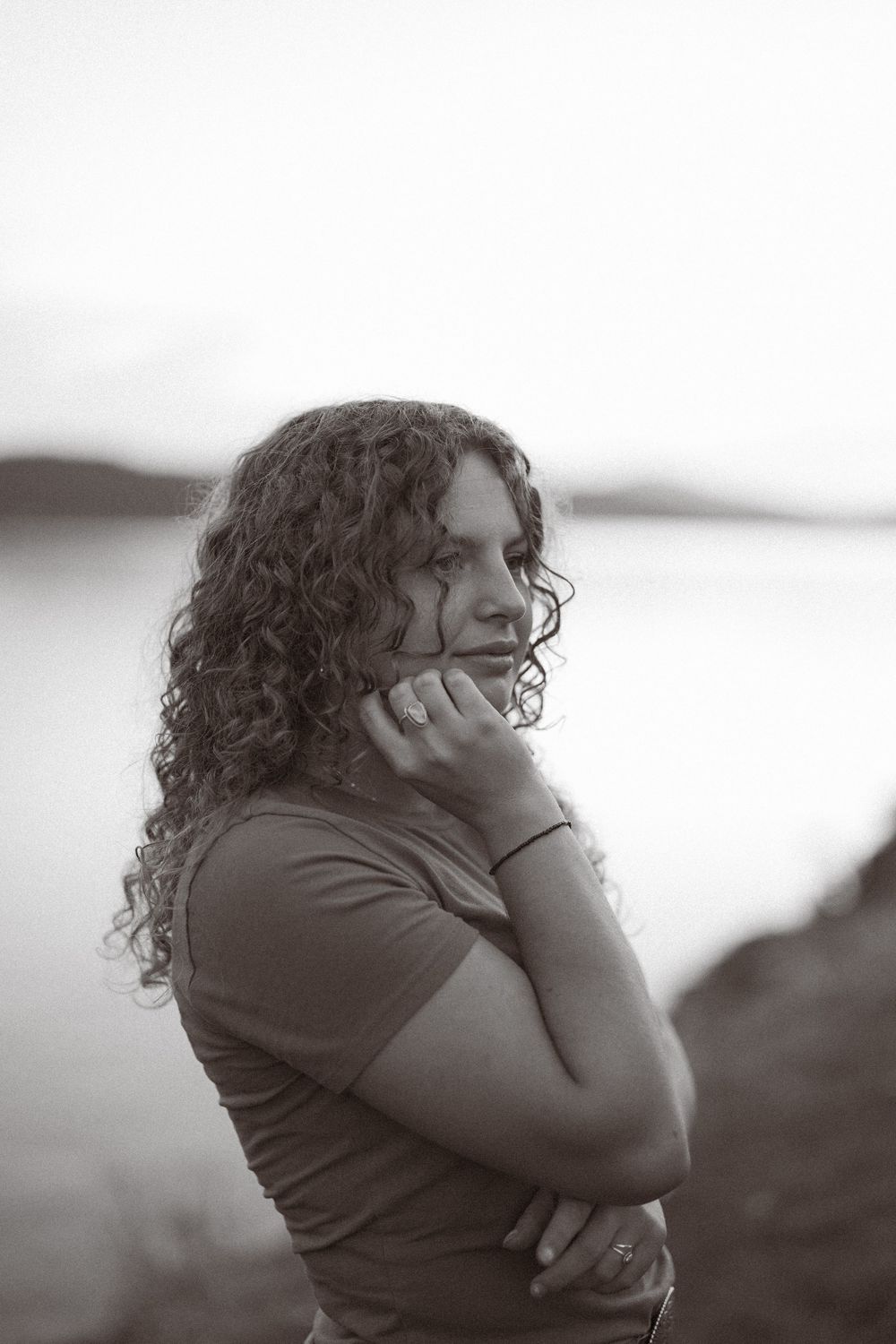 A person with long curly hair gazes thoughtfully into the distance in this black and white portrait photo near water.