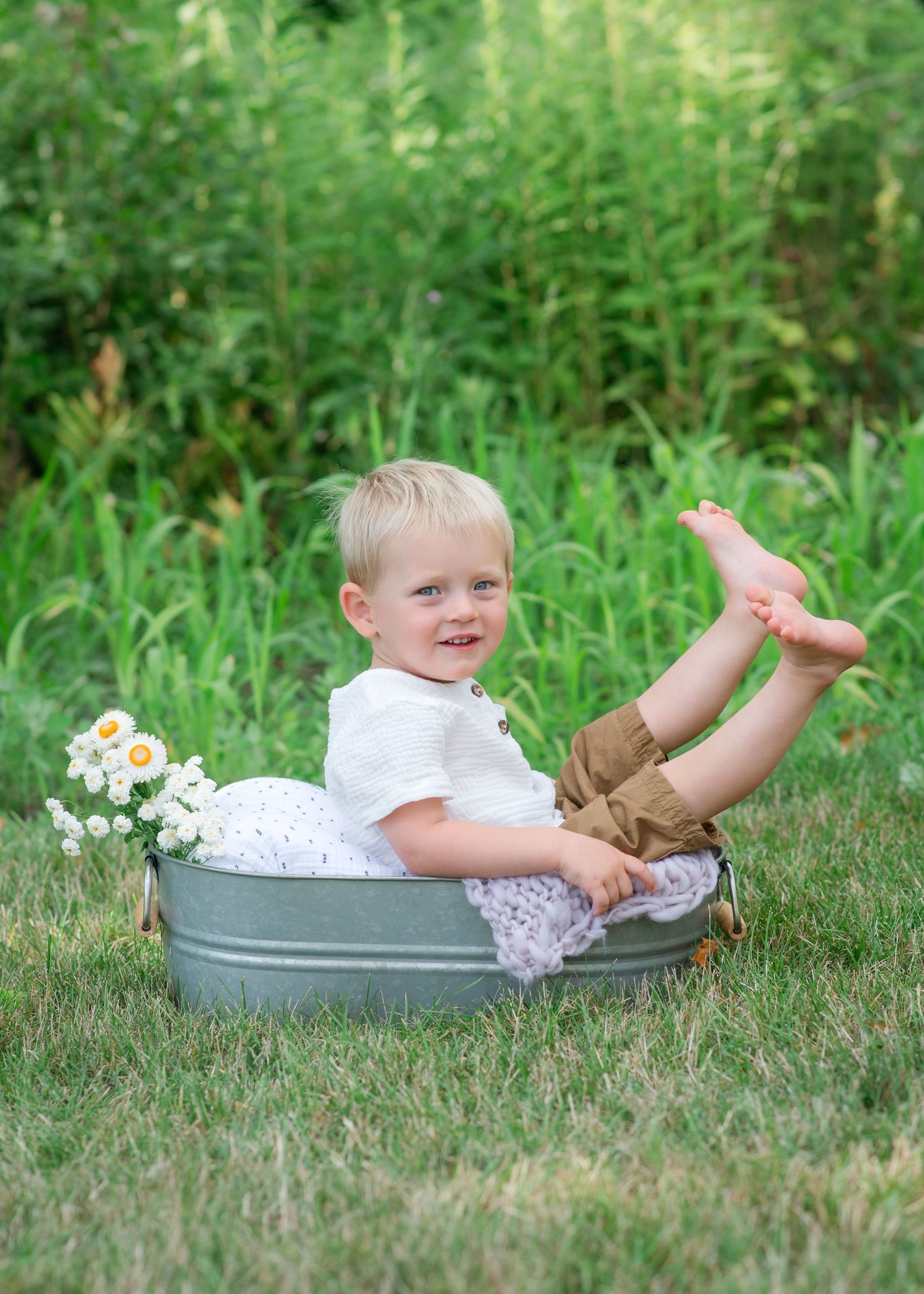 A child sits playfully in a galvanized metal tub outdoors with white flowers, surrounded by lush green grass and foliage.