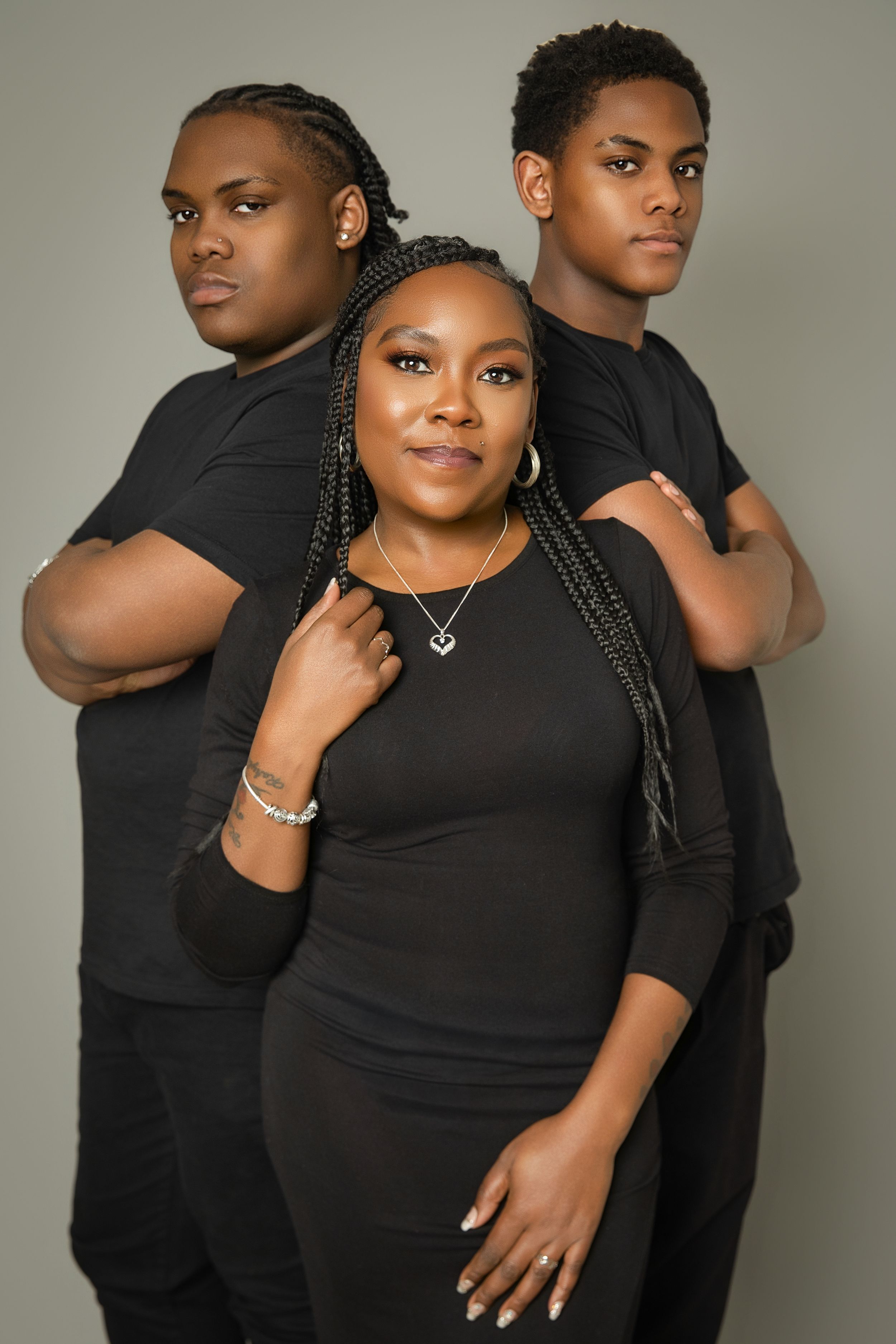 Three people in black outfits pose together for a stylish studio portrait against a neutral background.