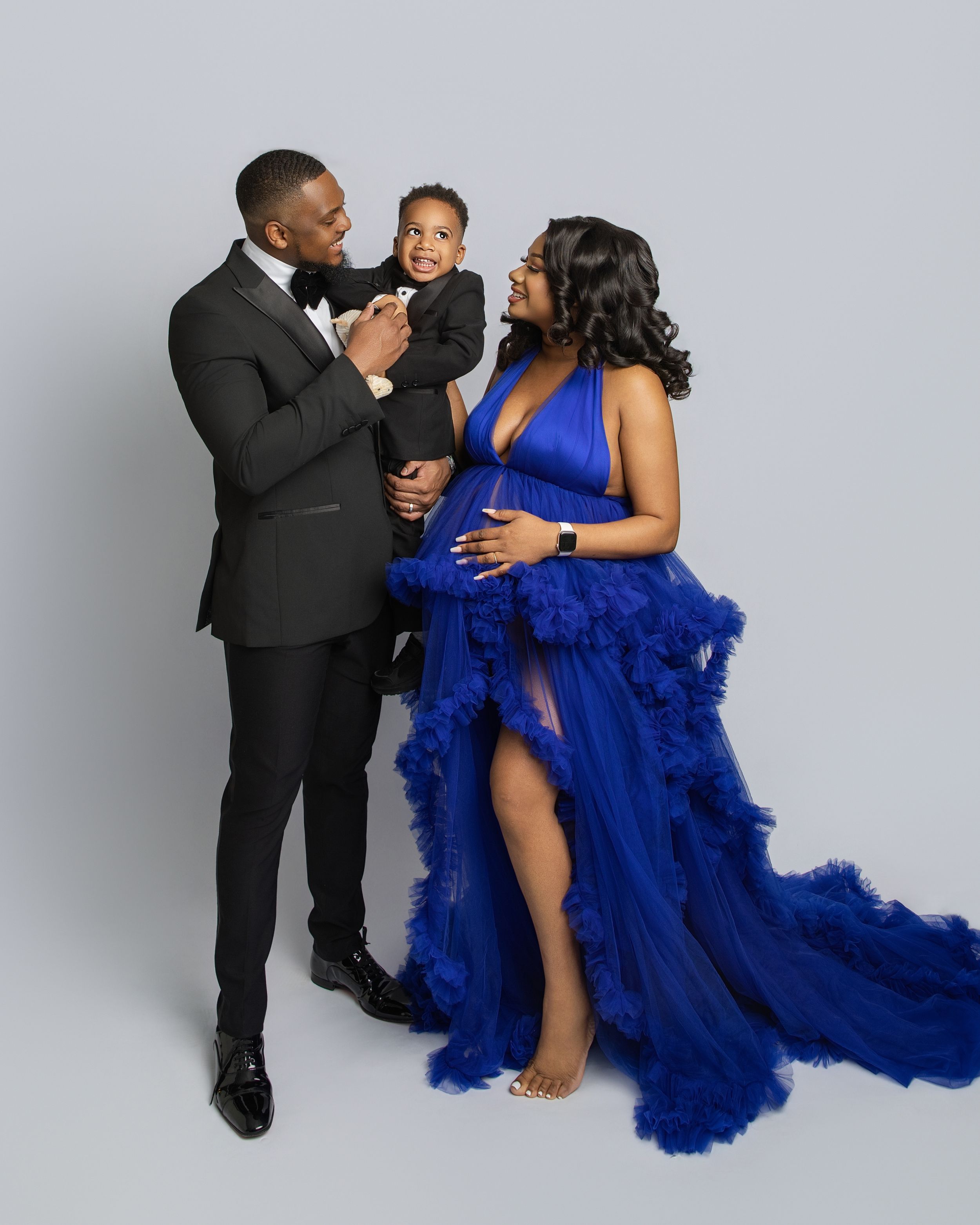 A couple in formal attire poses intimately against a white backdrop, with the woman wearing a flowing royal blue gown.