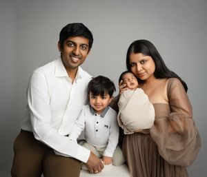 A family of four poses together in a studio setting wearing coordinated neutral colored outfits against a gray backdrop.
