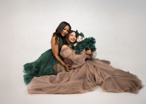 Two people pose together in flowing formal gowns of emerald green and taupe on a light backdrop.