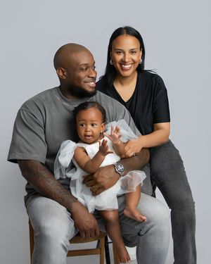 A happy family of three poses together for a portrait against a gray background while sitting on a wooden stool.