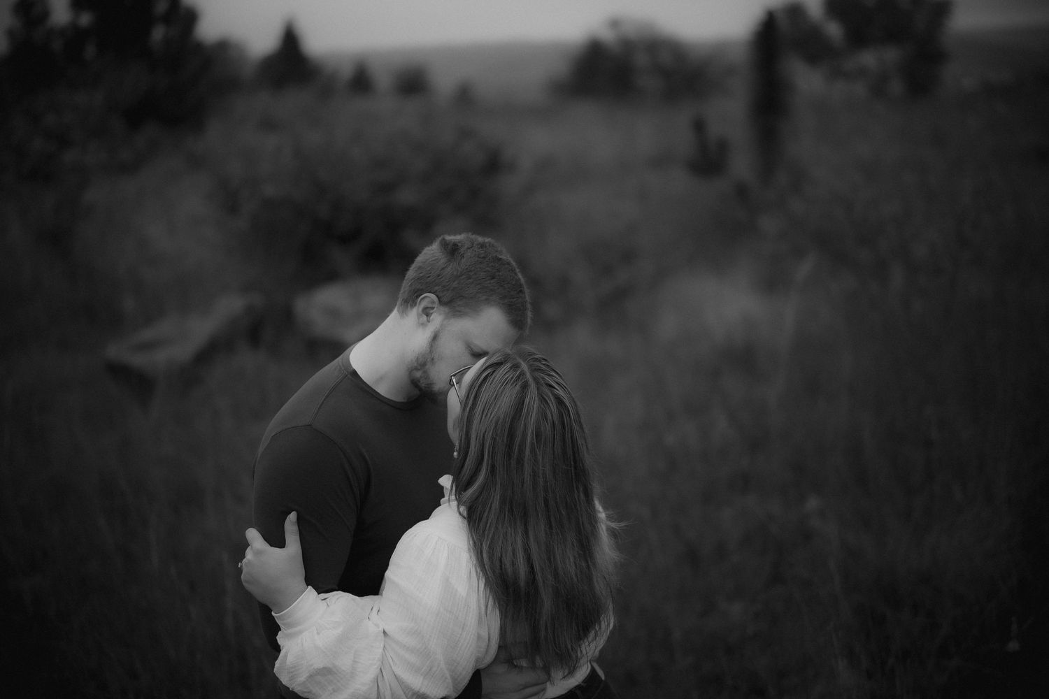 A black and white portrait of an embracing couple in a misty field at dusk.