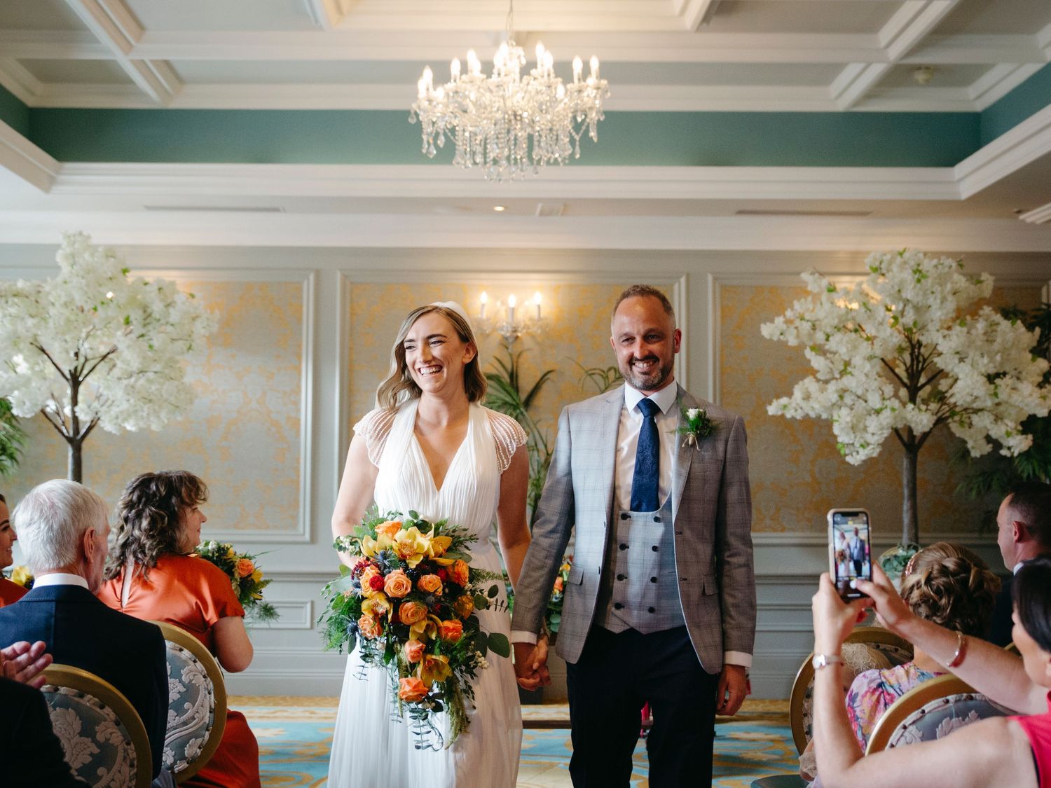 A couple walks down a grand aisle with elegant chandelier and floral decor during an indoor wedding ceremony.