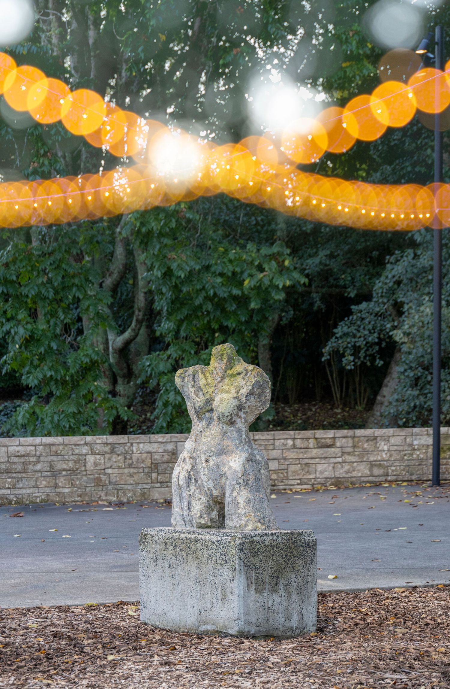 Moss-covered torso statue on a stone base, with orange string lights and a forested backdrop at dusk.