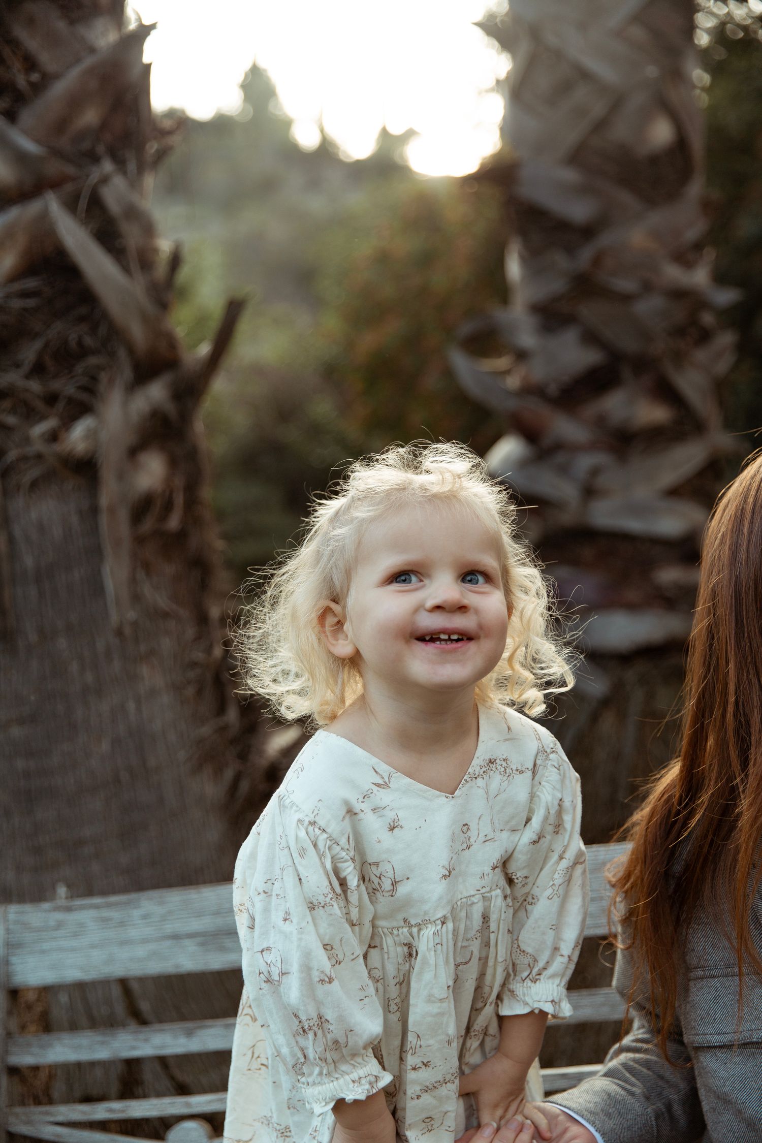 Child with curly blonde hair smiling while backlit by golden sunset among palm trees.